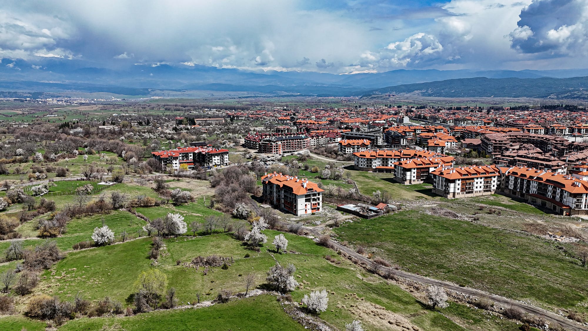 A wide aerial view of Bansko, Bulgaria, showing clusters of red-roofed apartment buildings set against green fields with blooming trees, stretching toward the distant Pirin Mountains under a partly cloudy sky