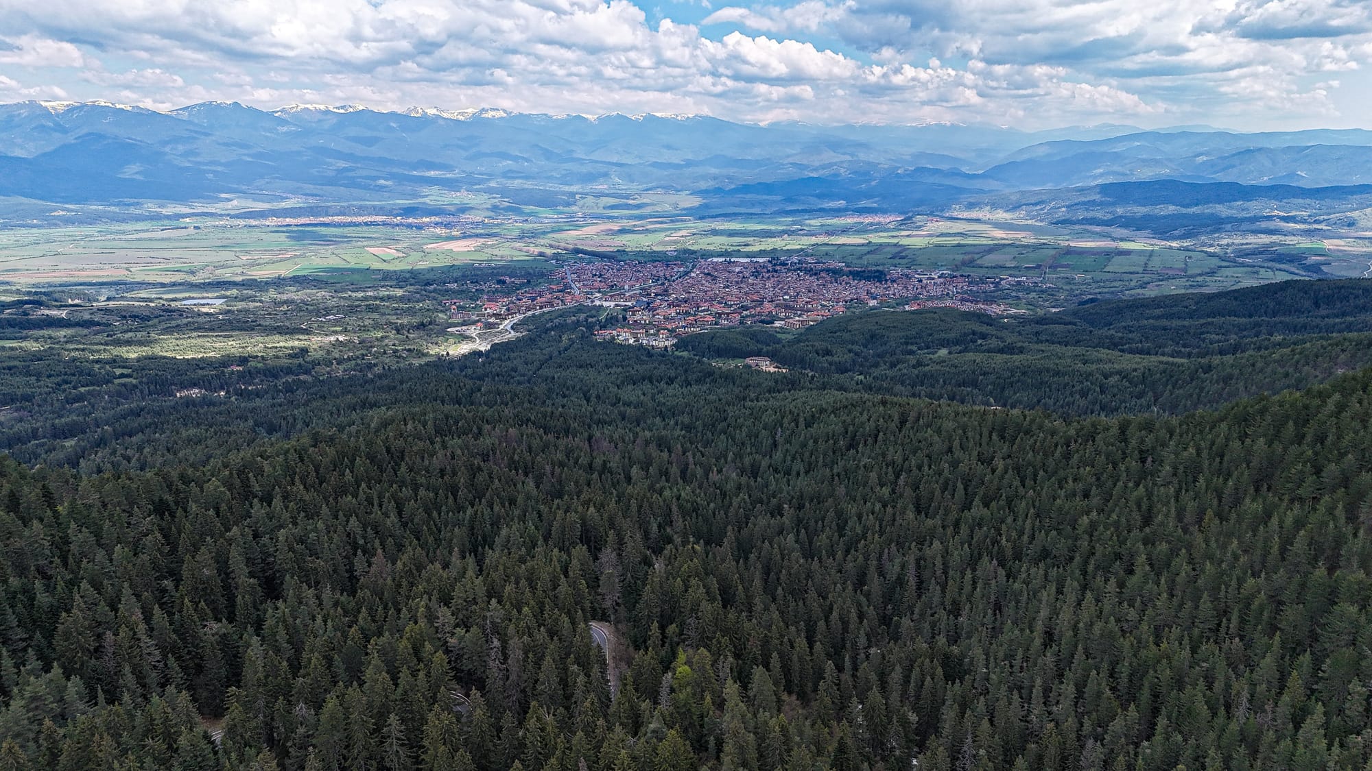 A panoramic view from above Bansko, showing the small mountain town in the valley with red-roofed buildings, surrounded by dense evergreen forests and distant layers of mountain ranges under a partly cloudy sky