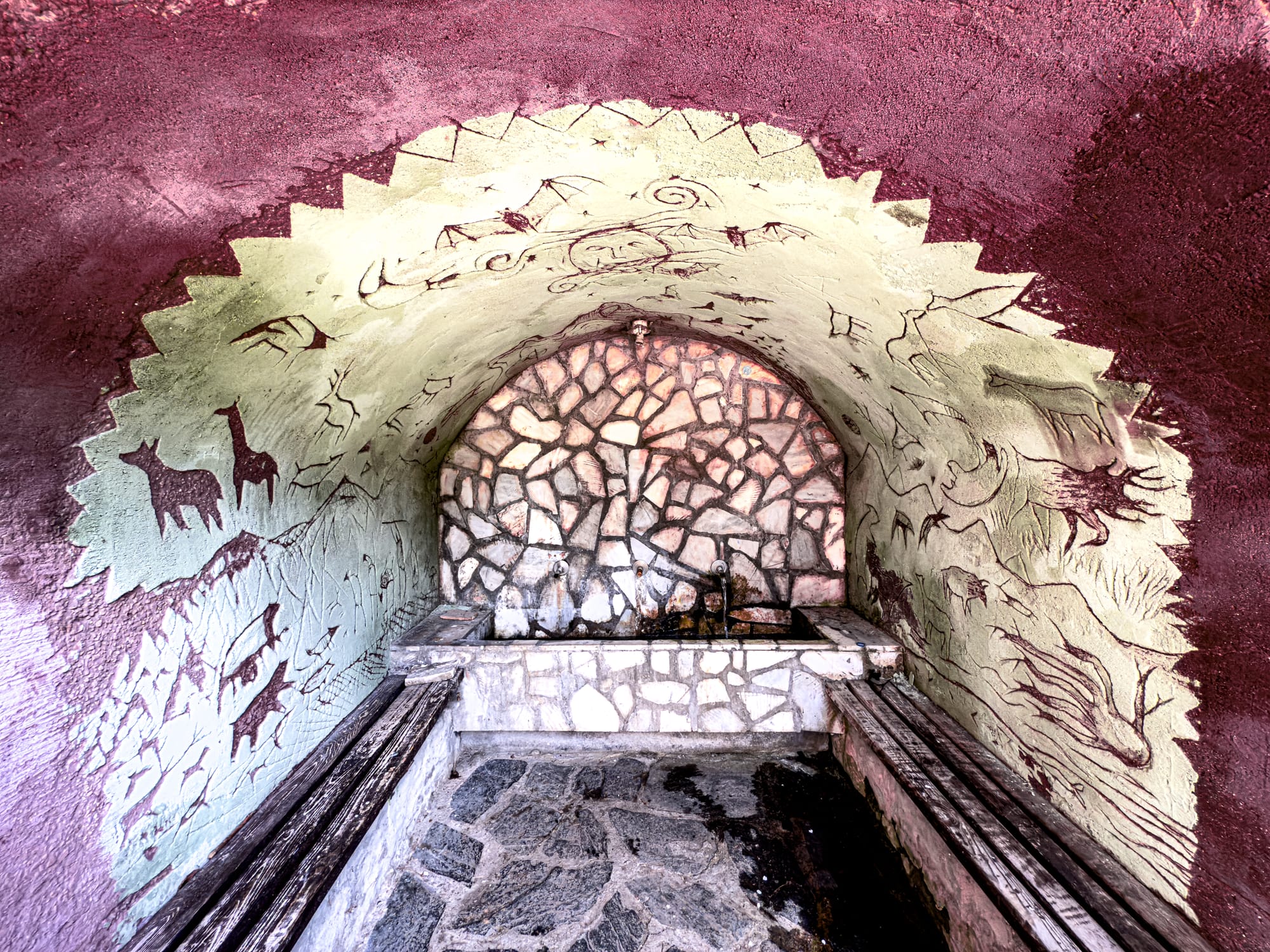 A stone water fountain alcove at The Secret Hotel near Bansko, decorated with cave-painting-style murals of deer, mountain goats, and abstract symbols on the curved walls, blending rustic benches and stone flooring into a mountain-inspired design