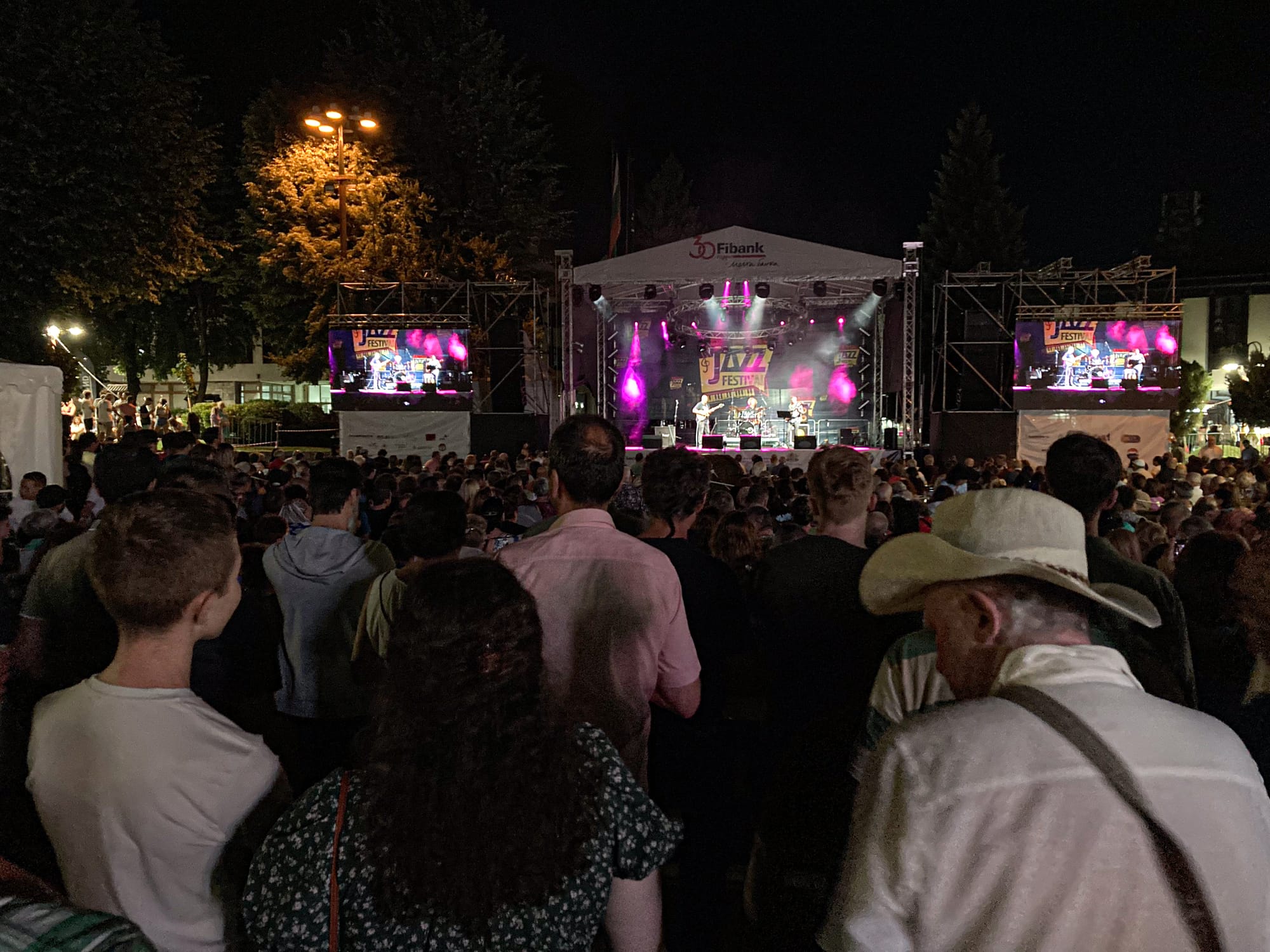 A large crowd gathered at night for the Bansko International Jazz Festival, with people watching musicians perform on a brightly lit outdoor stage under purple and pink lights, flanked by big screens displaying the live show