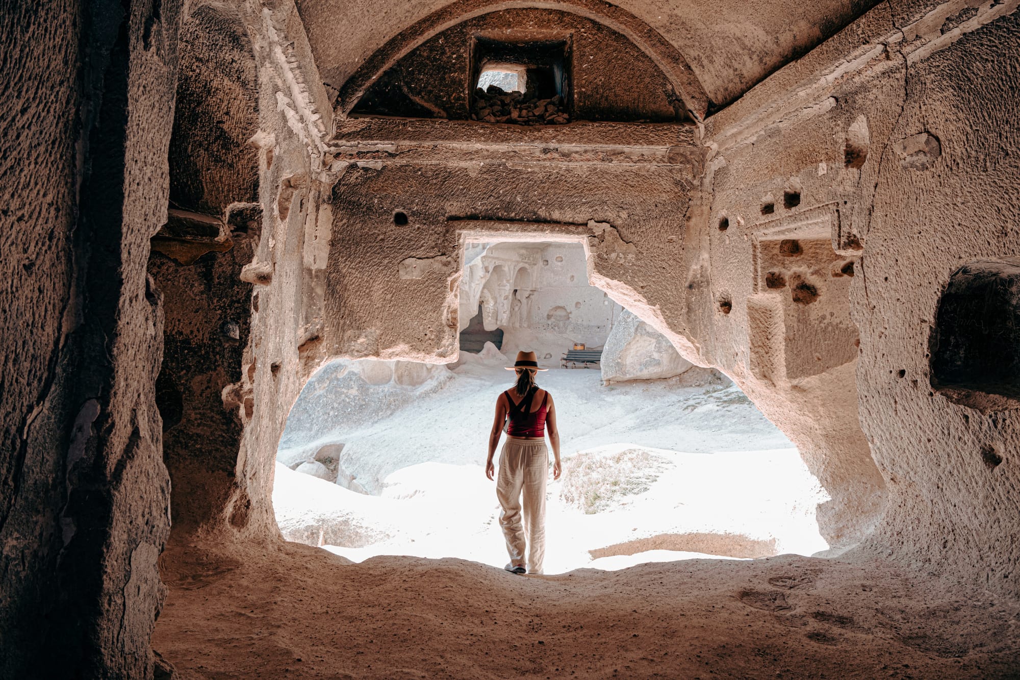 A person wearing a hat and light clothing stands at the entrance of a cave chamber inside Selime Monastery in Cappadocia, Turkey