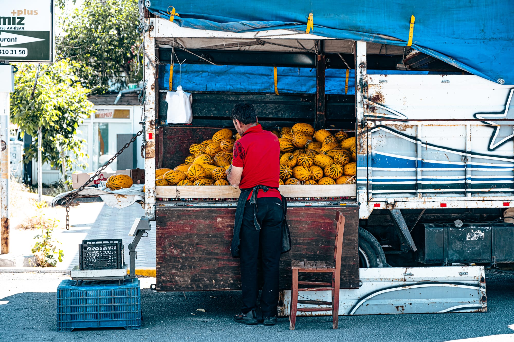 A man in a red shirt stands at the back of a truck in Göreme, Cappadocia, selling bright yellow striped melons piled high in wooden crates