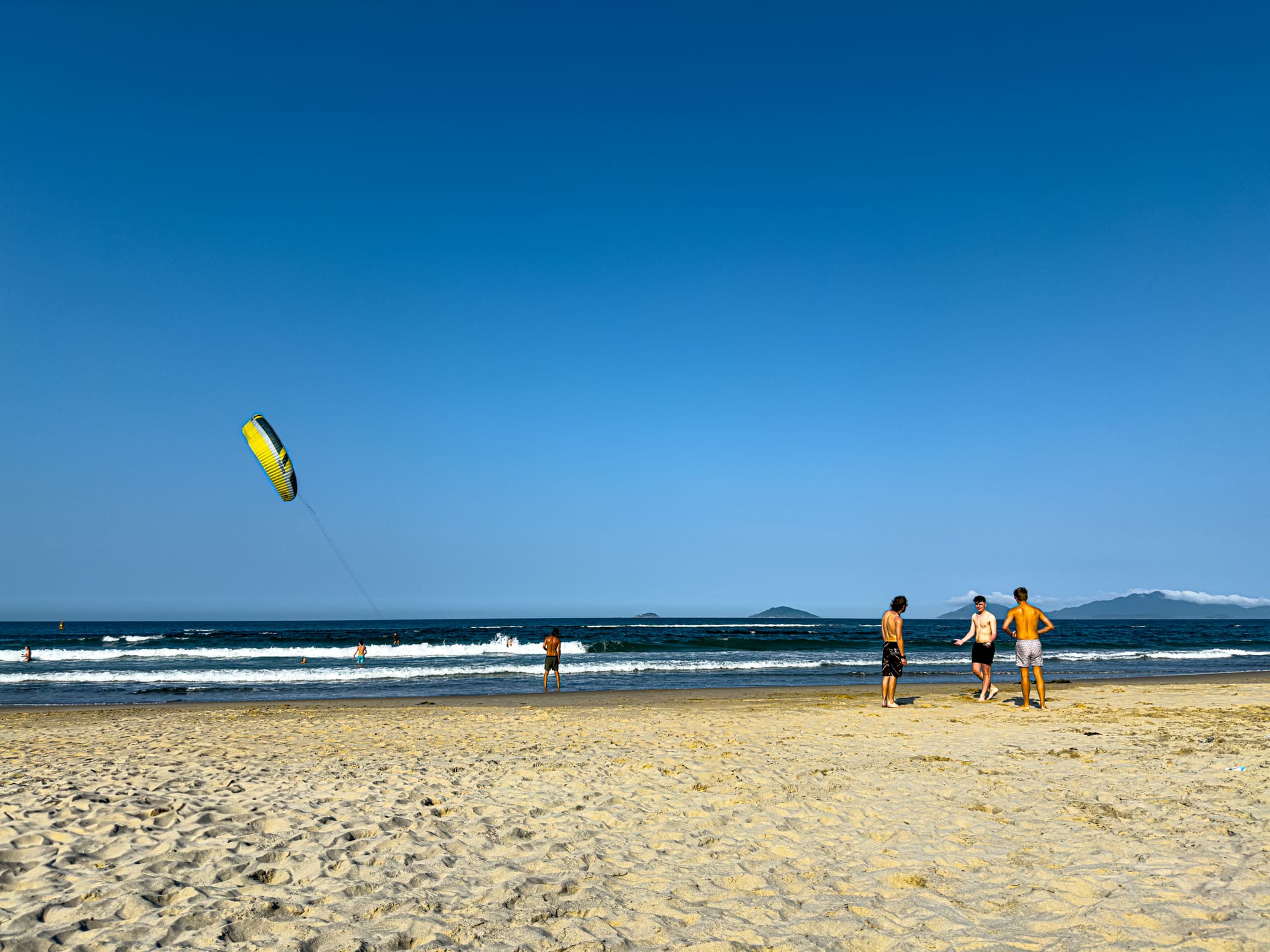 People relax on An Bang Beach in Hội An, with a kitesurfer’s yellow sail flying over the ocean and mountains visible on the horizon under a clear blue sky