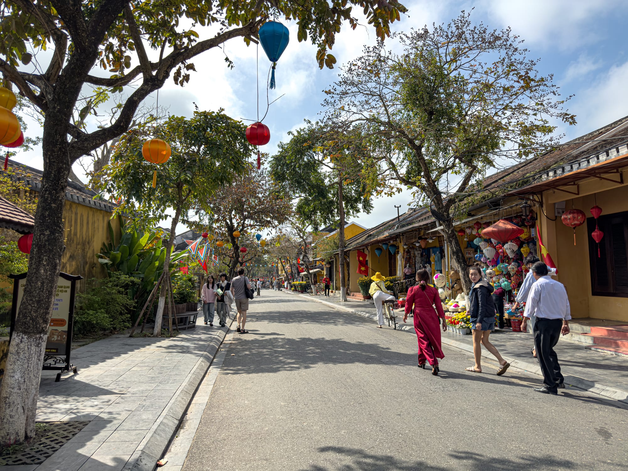 A lively street scene in Hội An’s Old Town showing people walking beneath hanging lanterns, with yellow-walled shops and market stalls along the shaded road