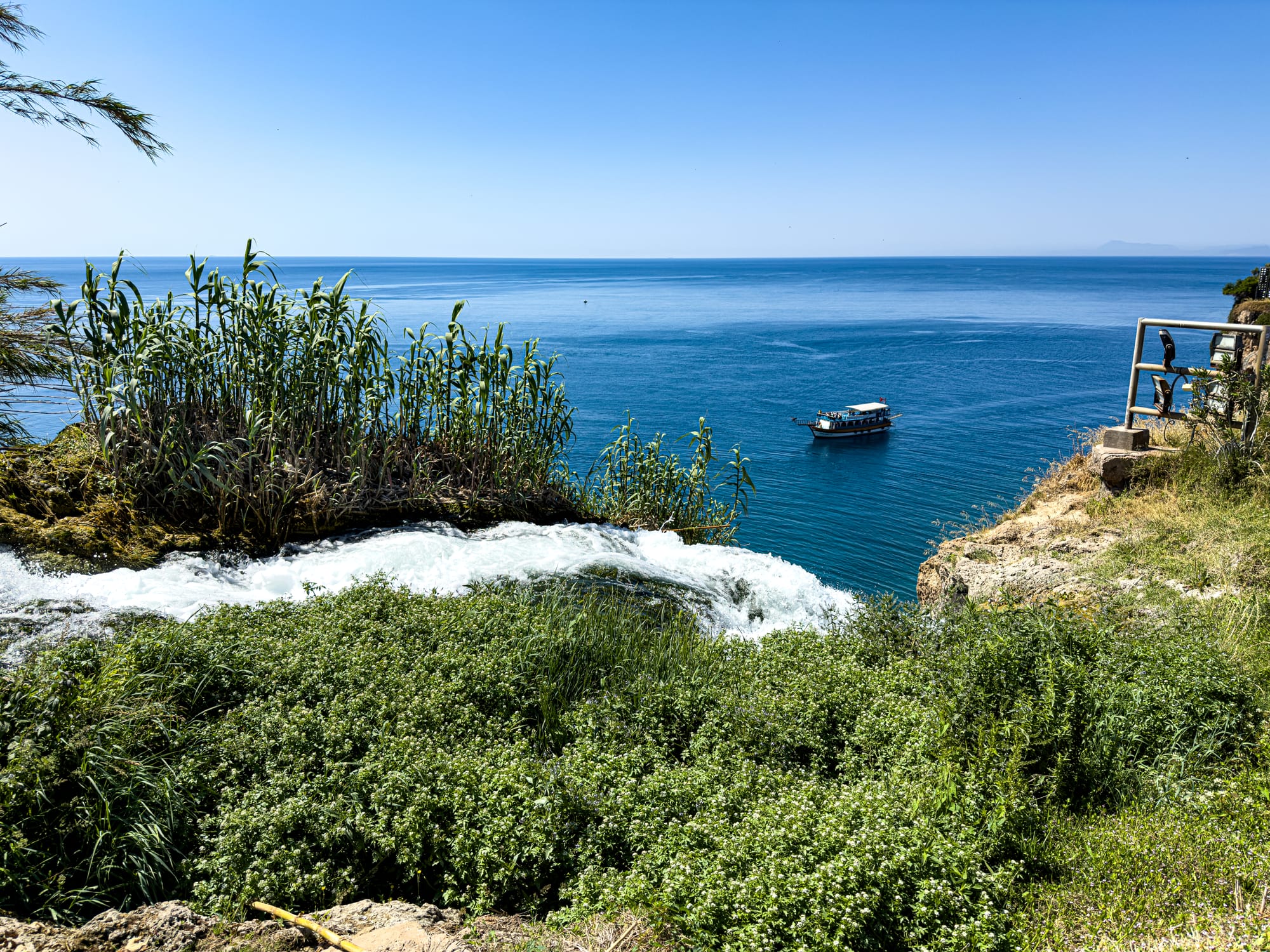 Overhead view of Düden Waterfall spilling toward the sea, with dense greenery in the foreground and a tour boat on the blue Mediterranean below