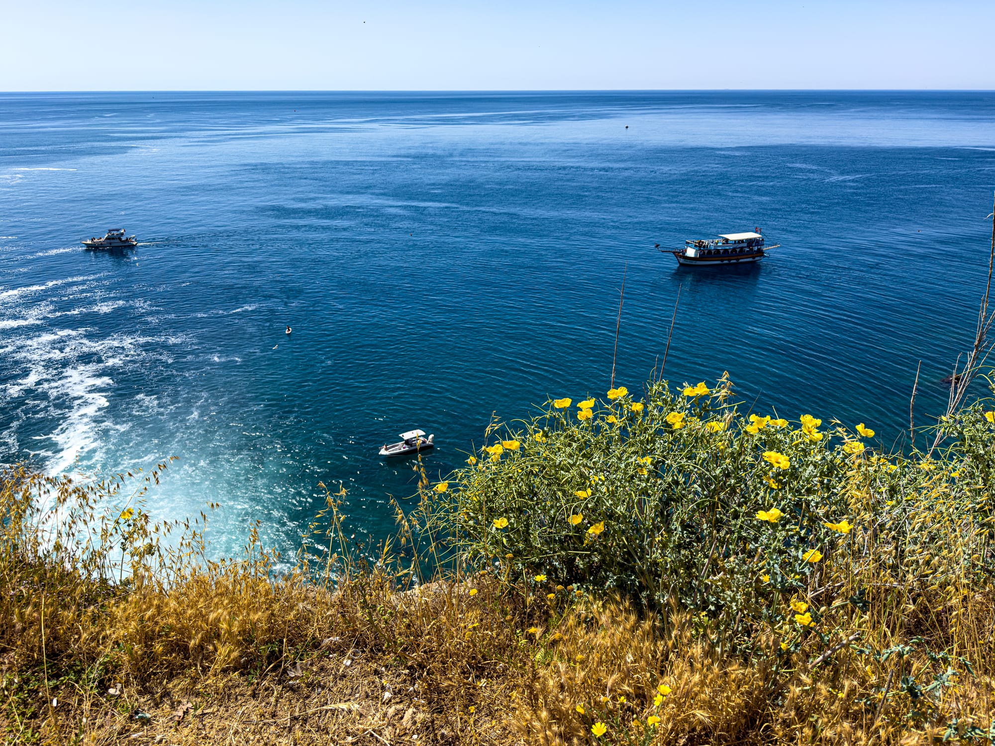 Boats on bright blue Mediterranean water seen from the cliffs near Düden Falls, with wild yellow flowers in the foreground