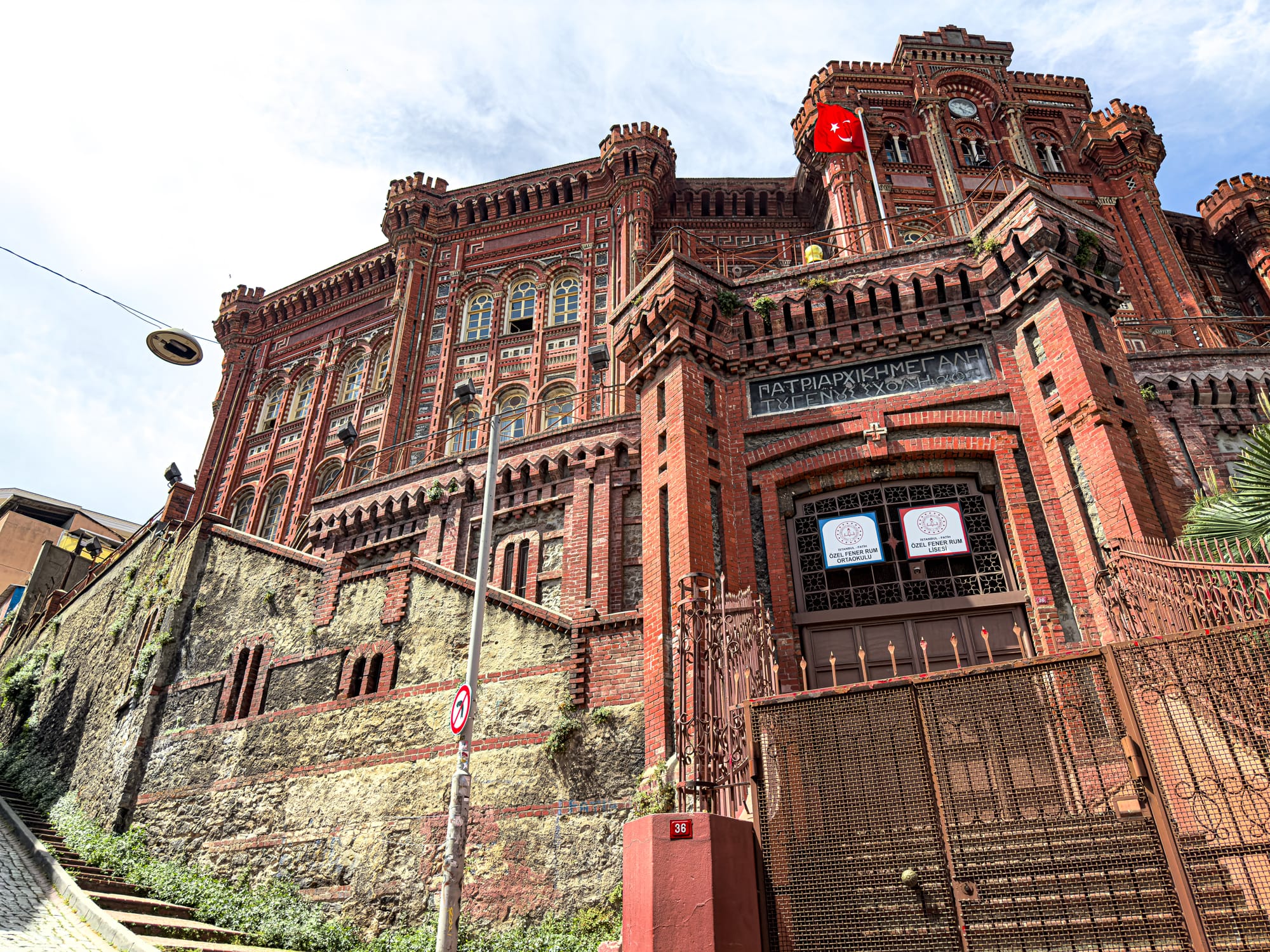 A towering red-brick structure with intricate details and a Turkish flag flying above—the historic Fener Greek High School sits elevated behind a stone wall and iron gate in Istanbul’s Fener district