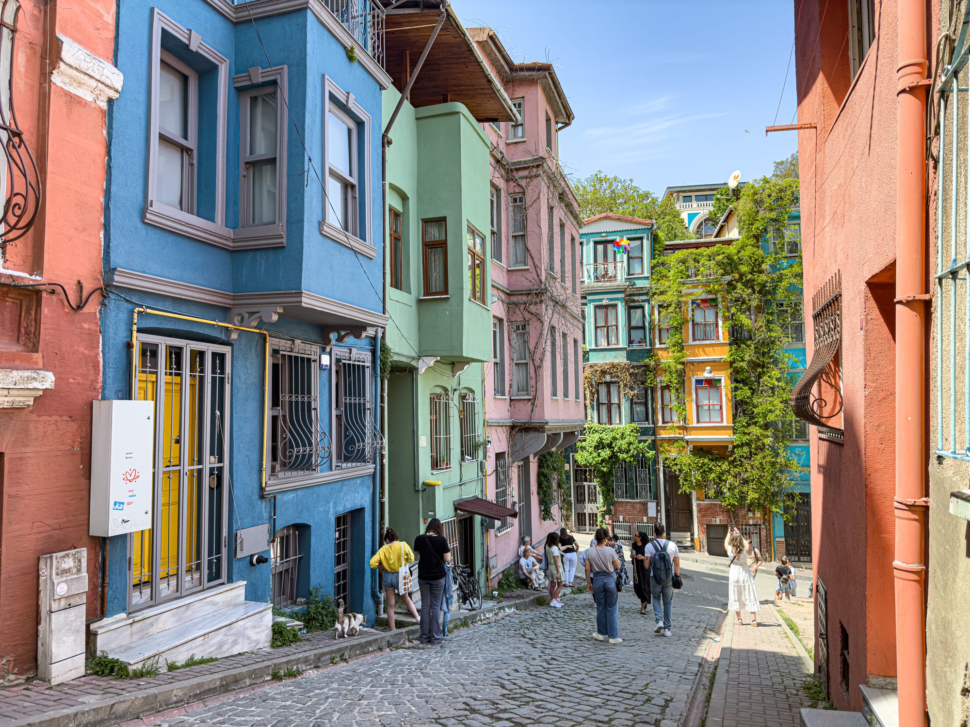 A cobbled street in Balat lined with colorful pastel buildings—blue, green, pink, and orange—with people walking and taking photos under bright afternoon sun