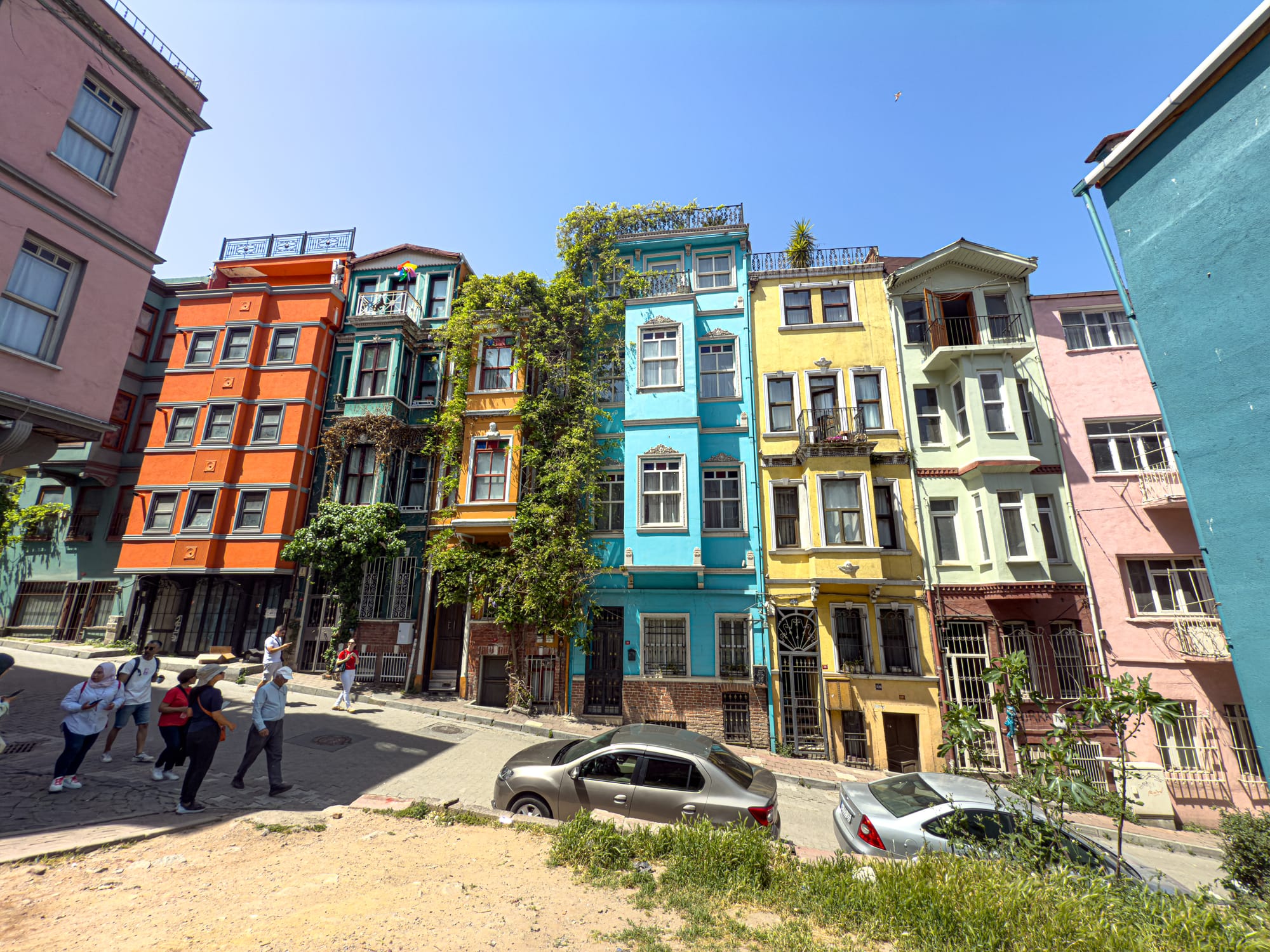 A sloped street in Balat lined with narrow, multicolored buildings—shades of orange, teal, yellow, pink, and blue—with people walking and cars parked in front