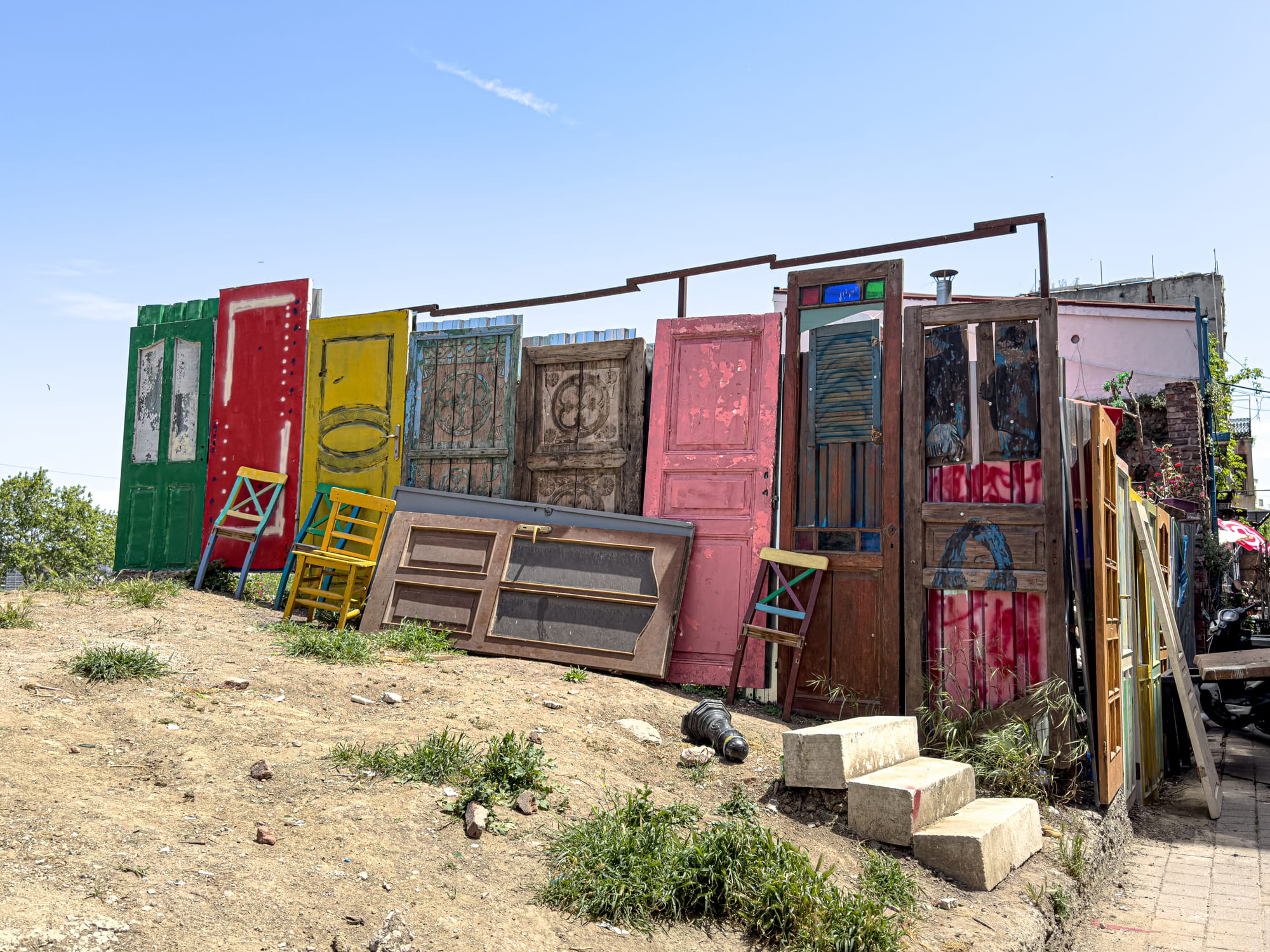 Colorful and weathered vintage doors arranged as a makeshift wall in a dirt clearing in Balat, surrounded by scattered chairs and blocks, showcasing the neighborhood’s playful, artistic spirit