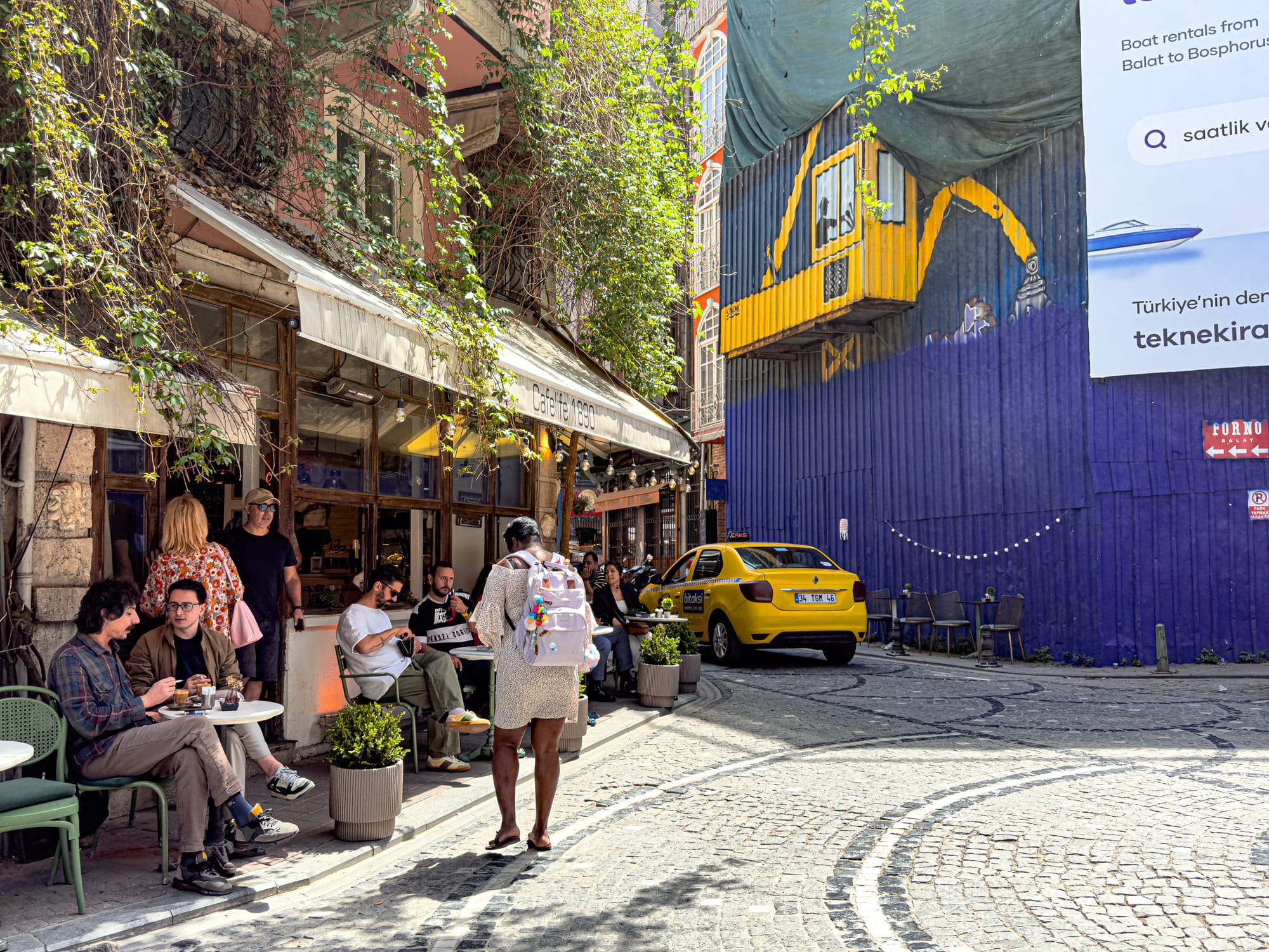 People sitting at outdoor tables along a café-lined street in Balat, Istanbul, with greenery, a yellow taxi, and colorful construction wall in the background