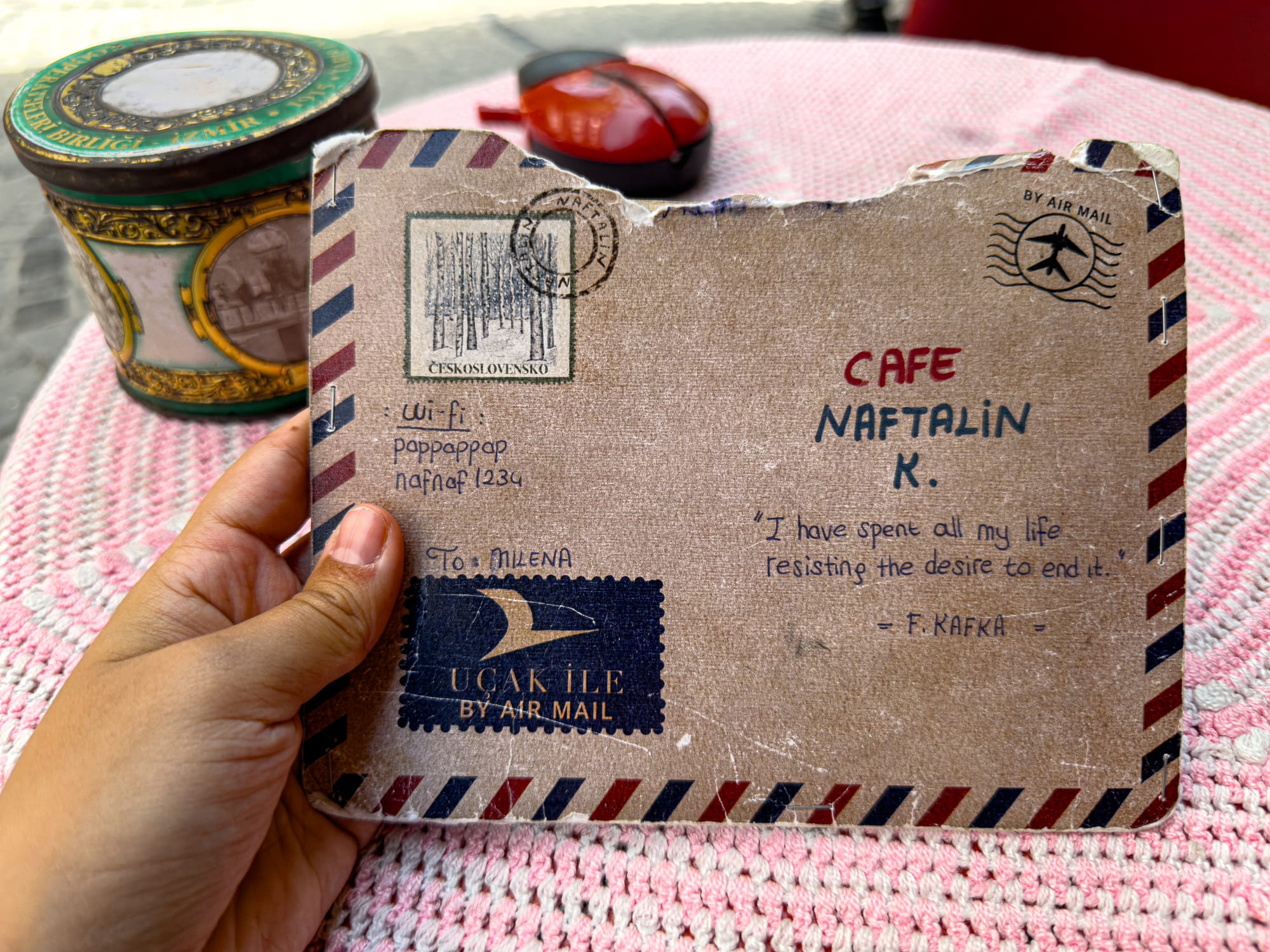 Close-up of a hand holding Cafe Naftalin’s creatively designed menu, made to look like a vintage airmail envelope with handwritten notes, stamps, and a Kafka quote, resting on a pink woven tablecloth