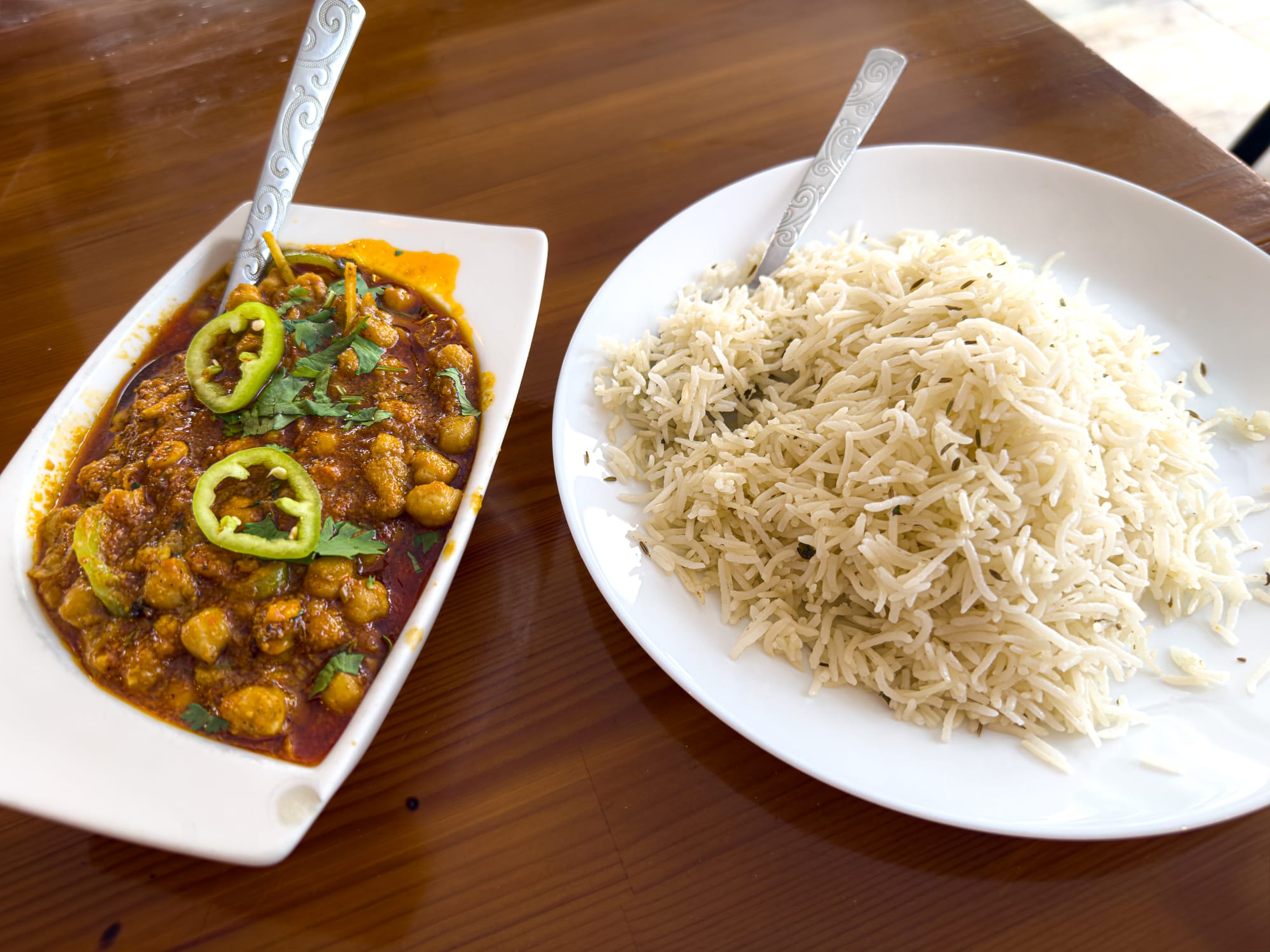Spicy chickpea curry (chana masala) garnished with green chili and cilantro, served alongside a heaping plate of plain basmati rice on a wooden table