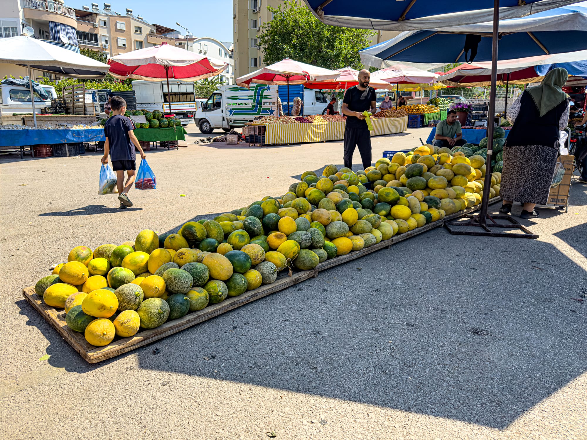 Melons stacked in rows at Antalya’s Friday Market with shoppers and umbrellas in the background