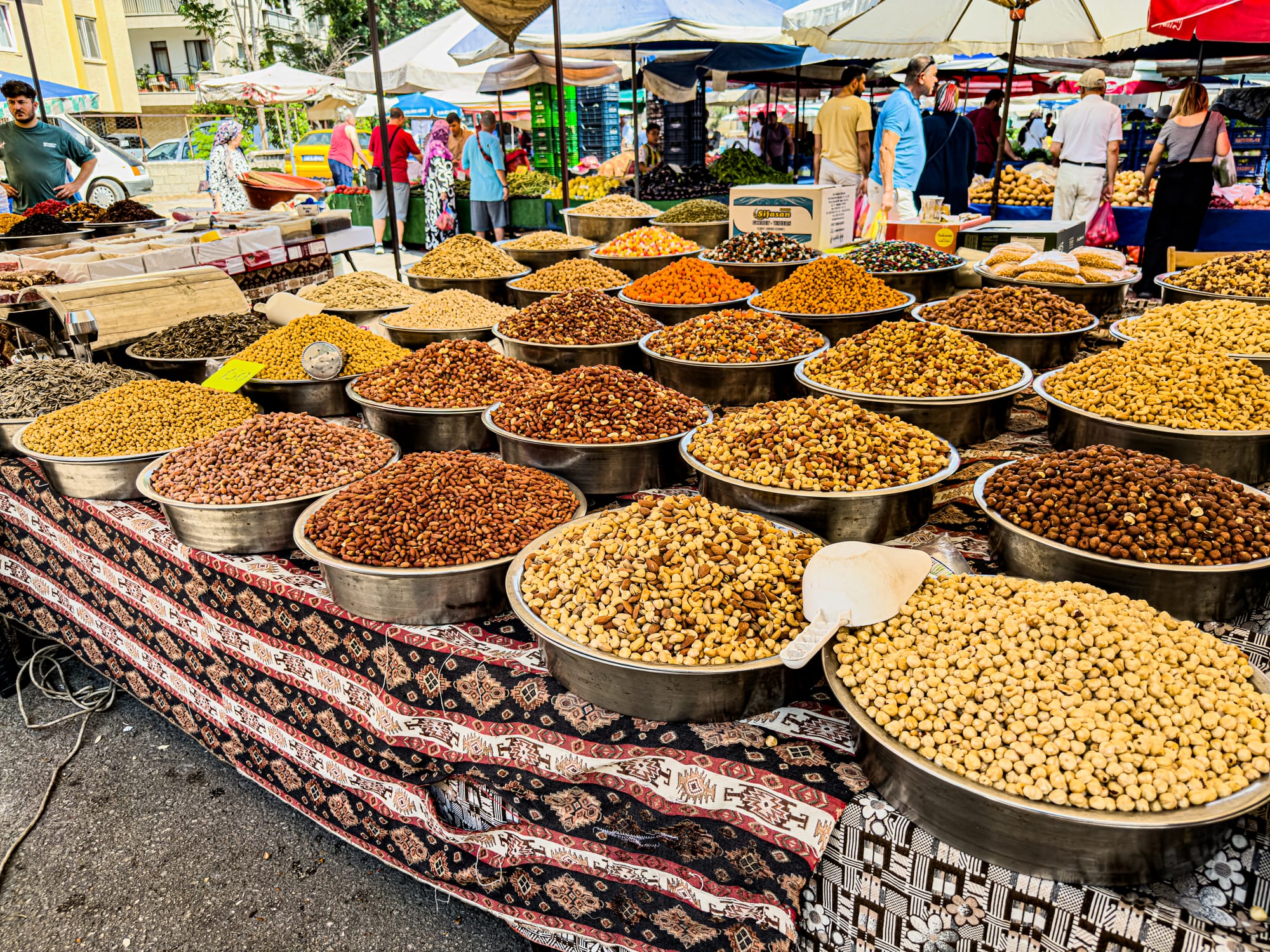 Metal bowls filled with assorted dried legumes, chickpeas, and nuts on patterned cloth at Antalya’s Friday Market