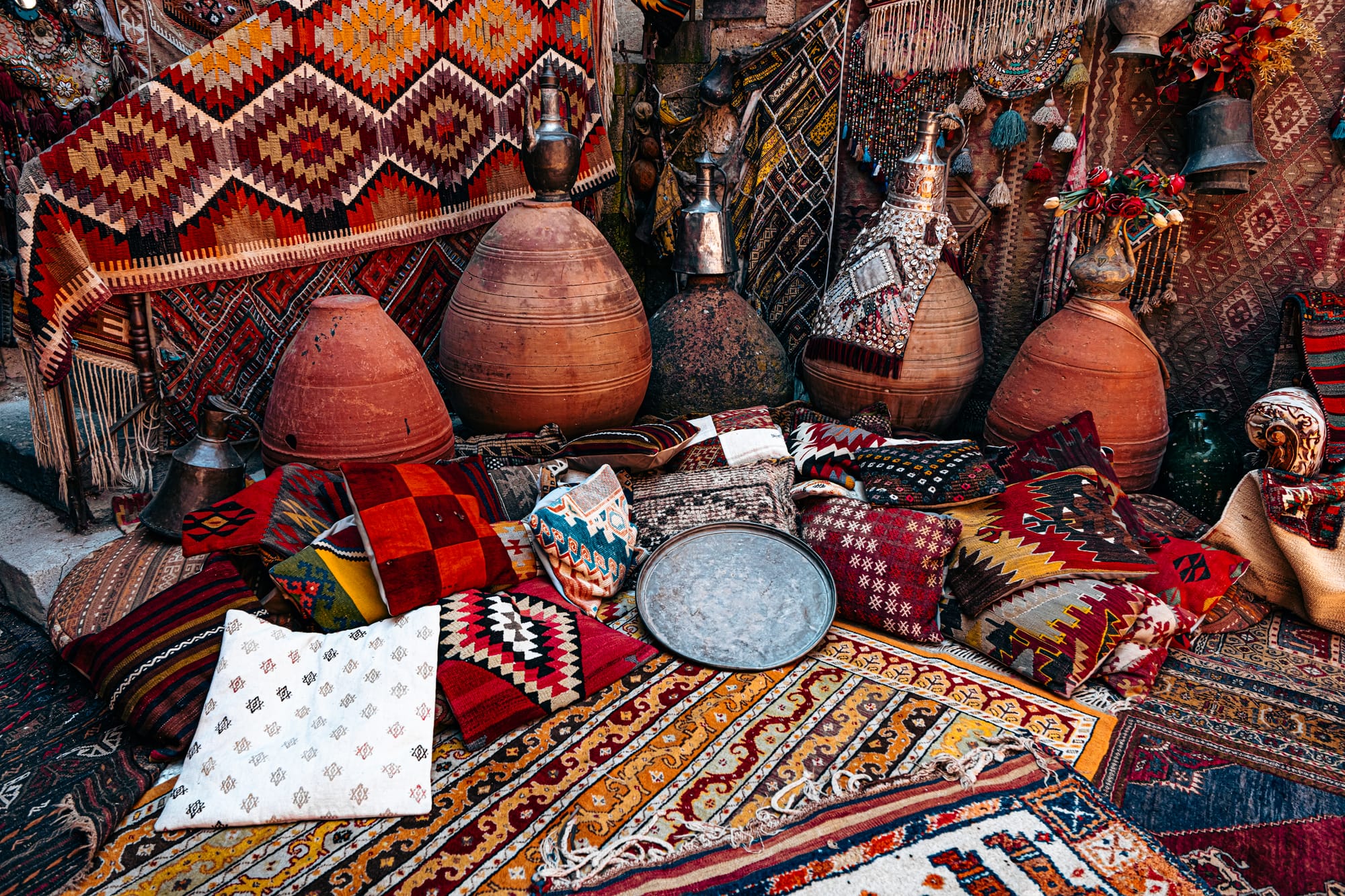 A display at Galerie İkman in Göreme featuring patterned rugs, colorful kilim pillows, large clay vessels with metal tops, woven wall hangings, and a round metal tray, arranged in a dense and vibrant composition