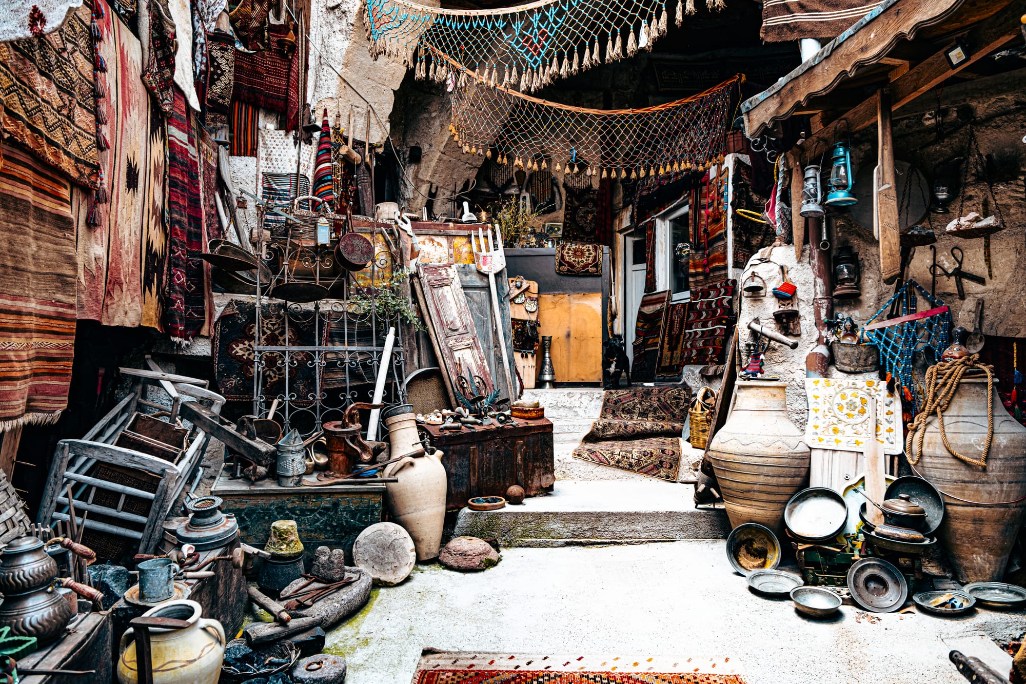 Entrance of the Cappadocia Cultural Antique Collection in Göreme, filled with an eclectic display of vintage artifacts including large clay jars, metal lanterns, wooden ladders, carpets, and old household tools arranged in a colorful, rustic courtyard