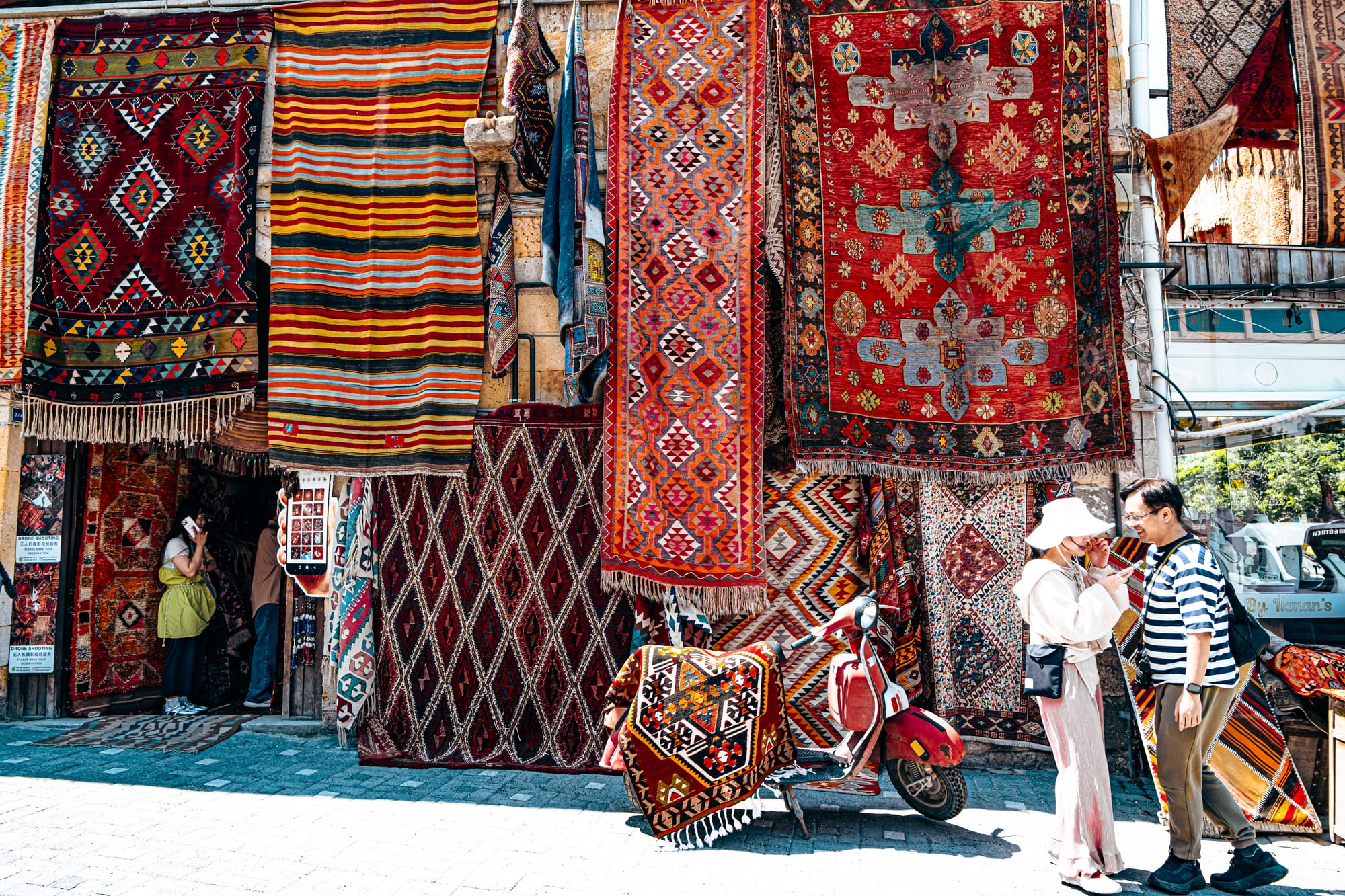 Exterior of Galerie İkman in Göreme, with vibrant Turkish carpets hanging above a red scooter and visitors browsing outside