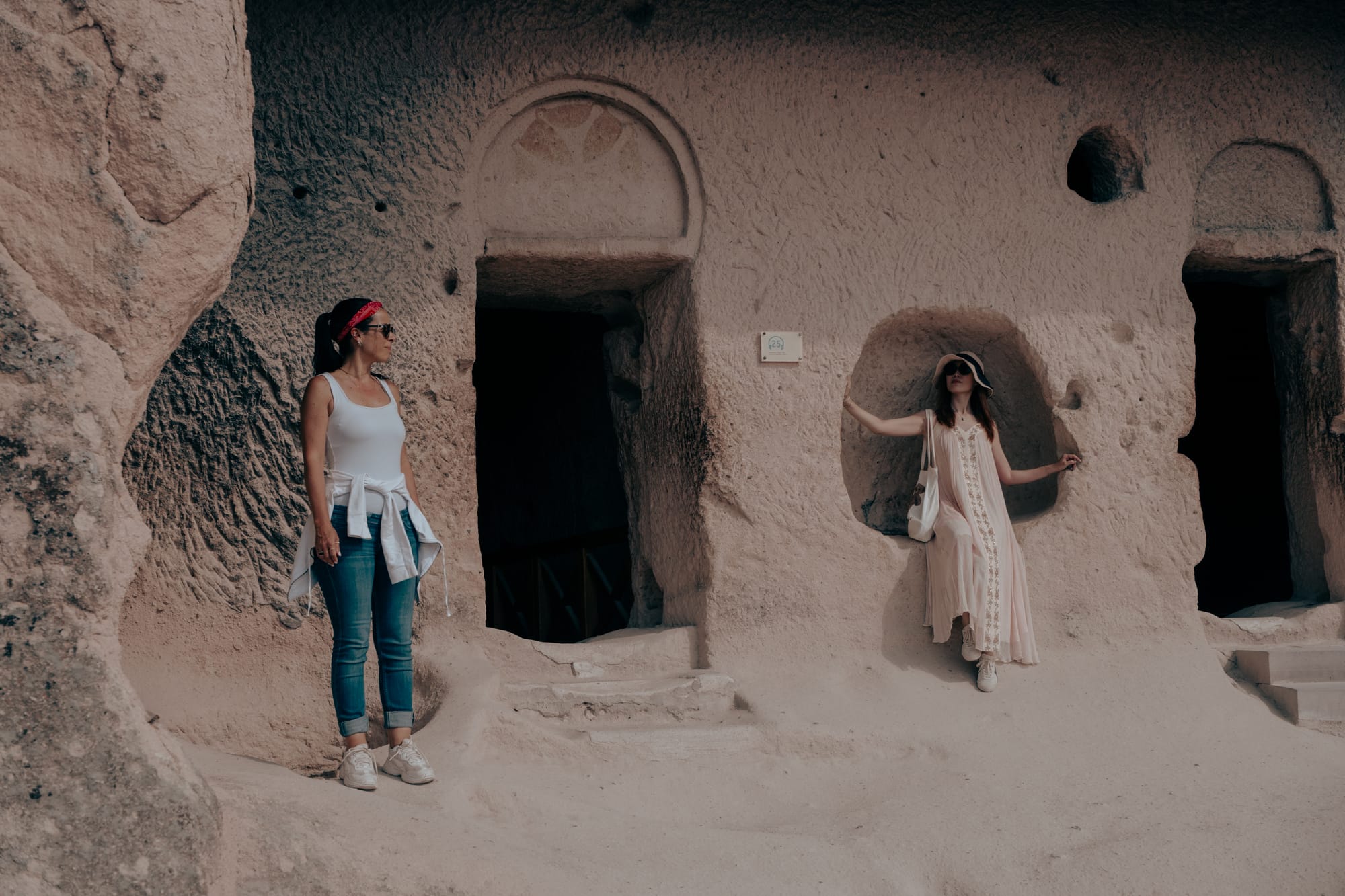 Two women stand outside carved rock entrances at Göreme Open Air Museum, one leaning in a niche and the other standing near a doorway