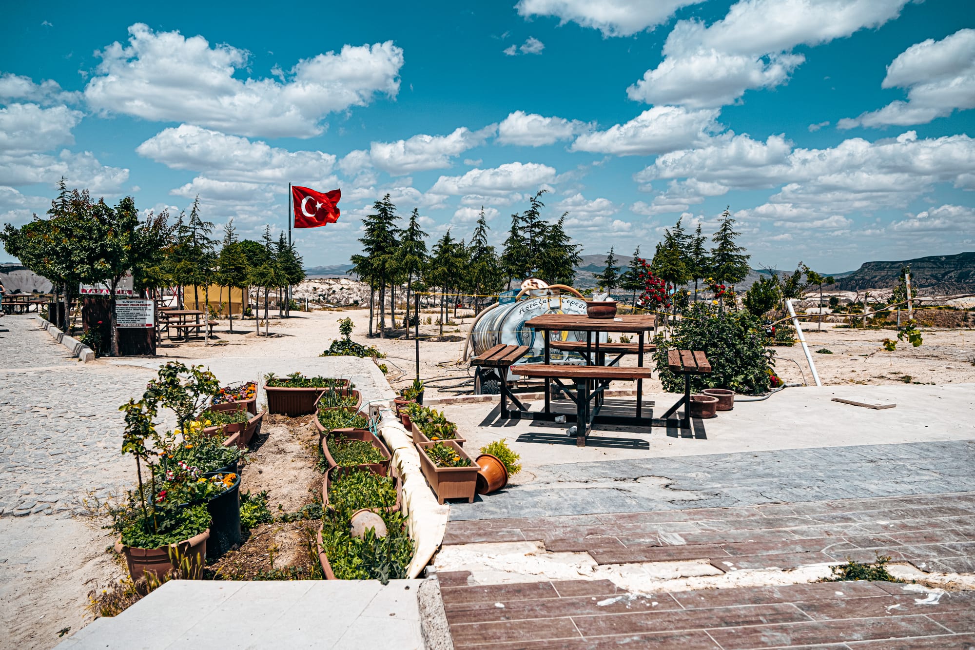 Outdoor seating area at Göreme Panorama viewpoint with picnic tables, potted plants, a Turkish flag waving in the background, and trees under a bright blue sky with scattered clouds