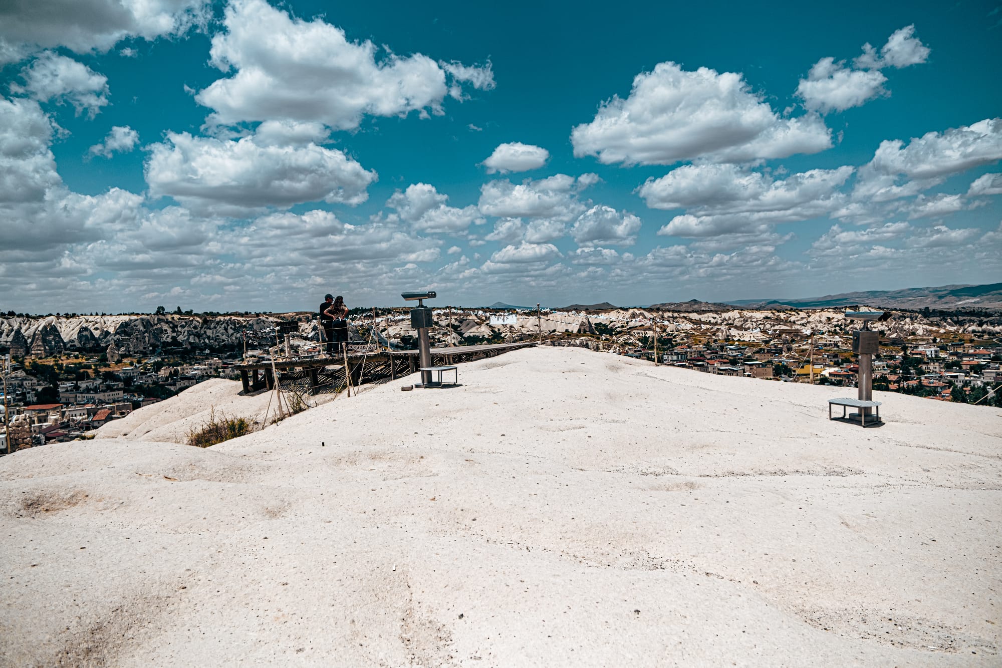 Wide view of the Göreme Panorama viewpoint showing telescopes and visitors overlooking Cappadocia’s valleys and rock formations on a sunny day with a bright blue sky and scattered clouds
