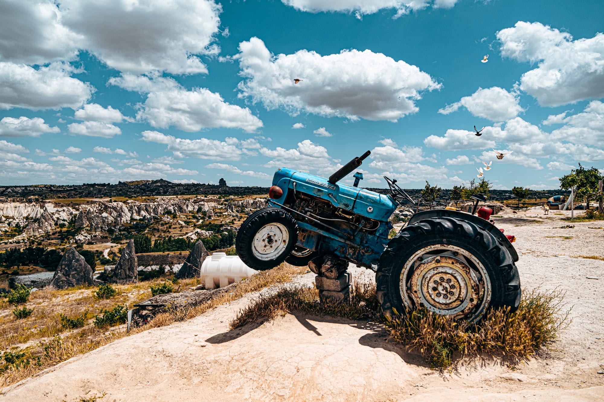 A blue tractor balanced on its rear wheels atop a rocky ledge at Göreme Panorama, with Cappadocia’s fairy chimneys and valleys stretching into the distance under a bright blue sky with scattered clouds
