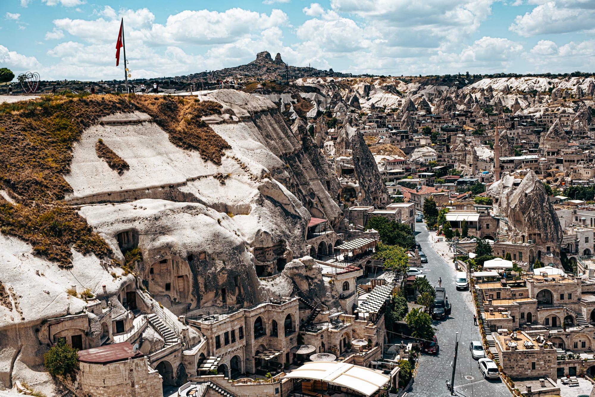 Elevated view of Göreme town’s cave hotels and rock formations, with the Turkish flag flying at the Panorama viewpoint and Uçhisar Castle visible on the horizon