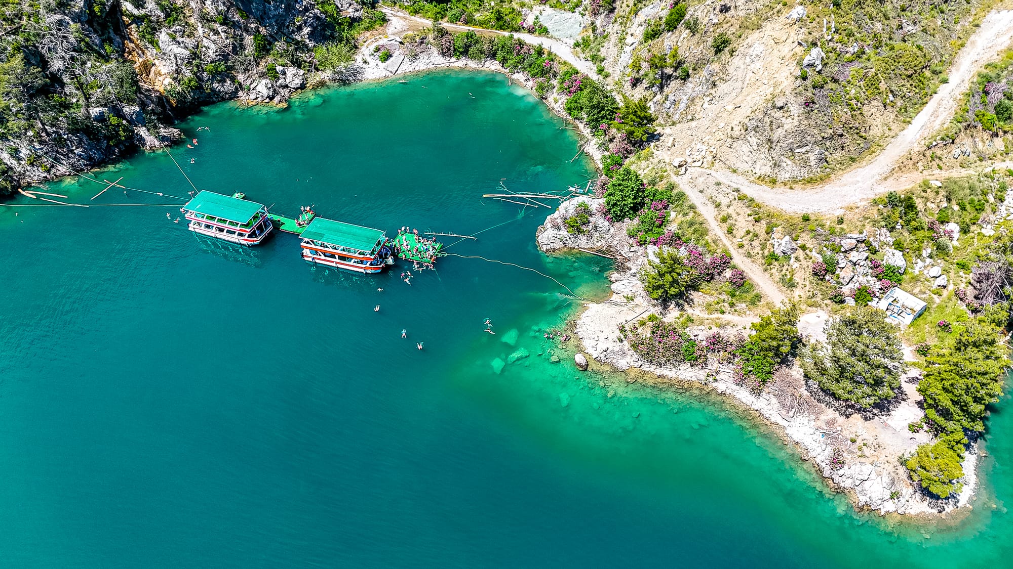Aerial view of a swimming cove in Green Canyon with turquoise water, anchored boats, and people swimming near the shore surrounded by rocky cliffs and blooming pink oleander