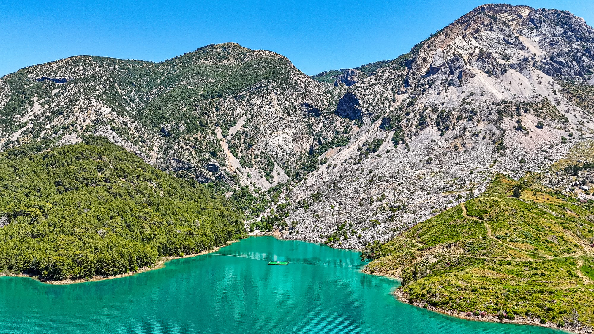 A boat cruising through turquoise water surrounded by steep forested hills and rocky mountains in the Green Canyon of Turkey