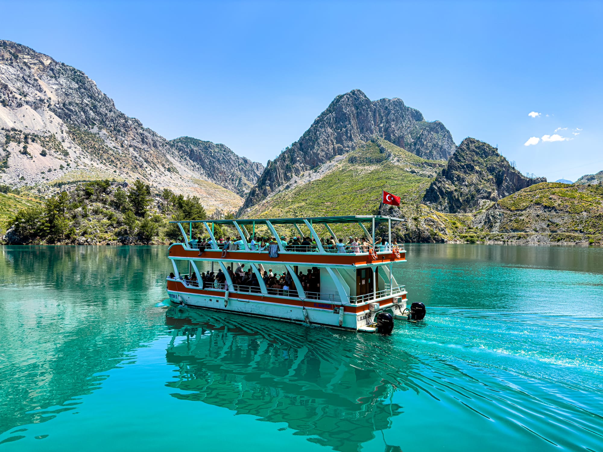 A two-level tour boat with a Turkish flag glides through the vibrant turquoise waters of Green Canyon, framed by steep, forested mountains and clear blue skies
