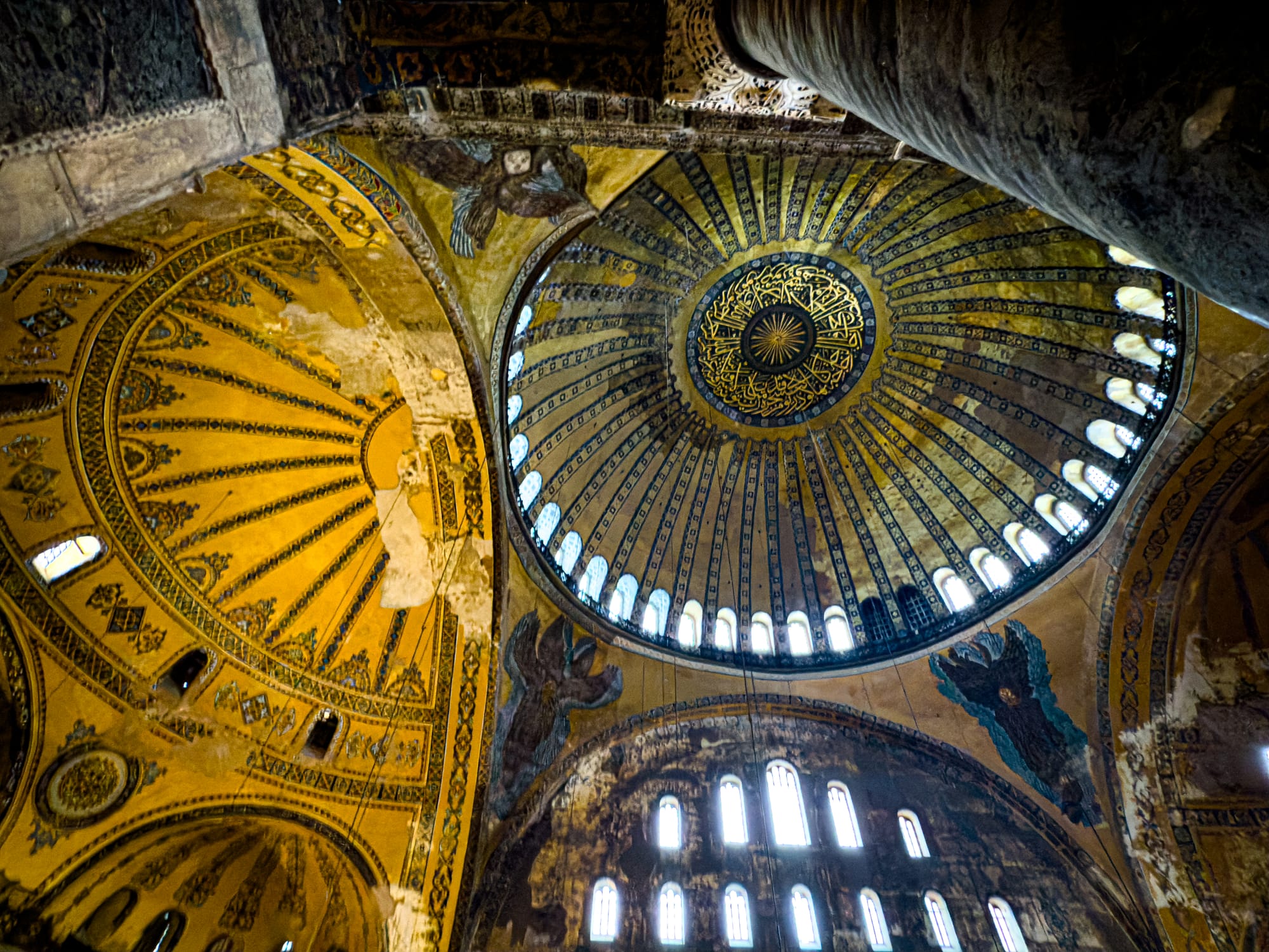 Interior view of the Hagia Sophia’s dome showing a blend of Byzantine angelic figures (seraphim), Arabic calligraphy, and golden-yellow geometric ornamentation illuminated by natural light