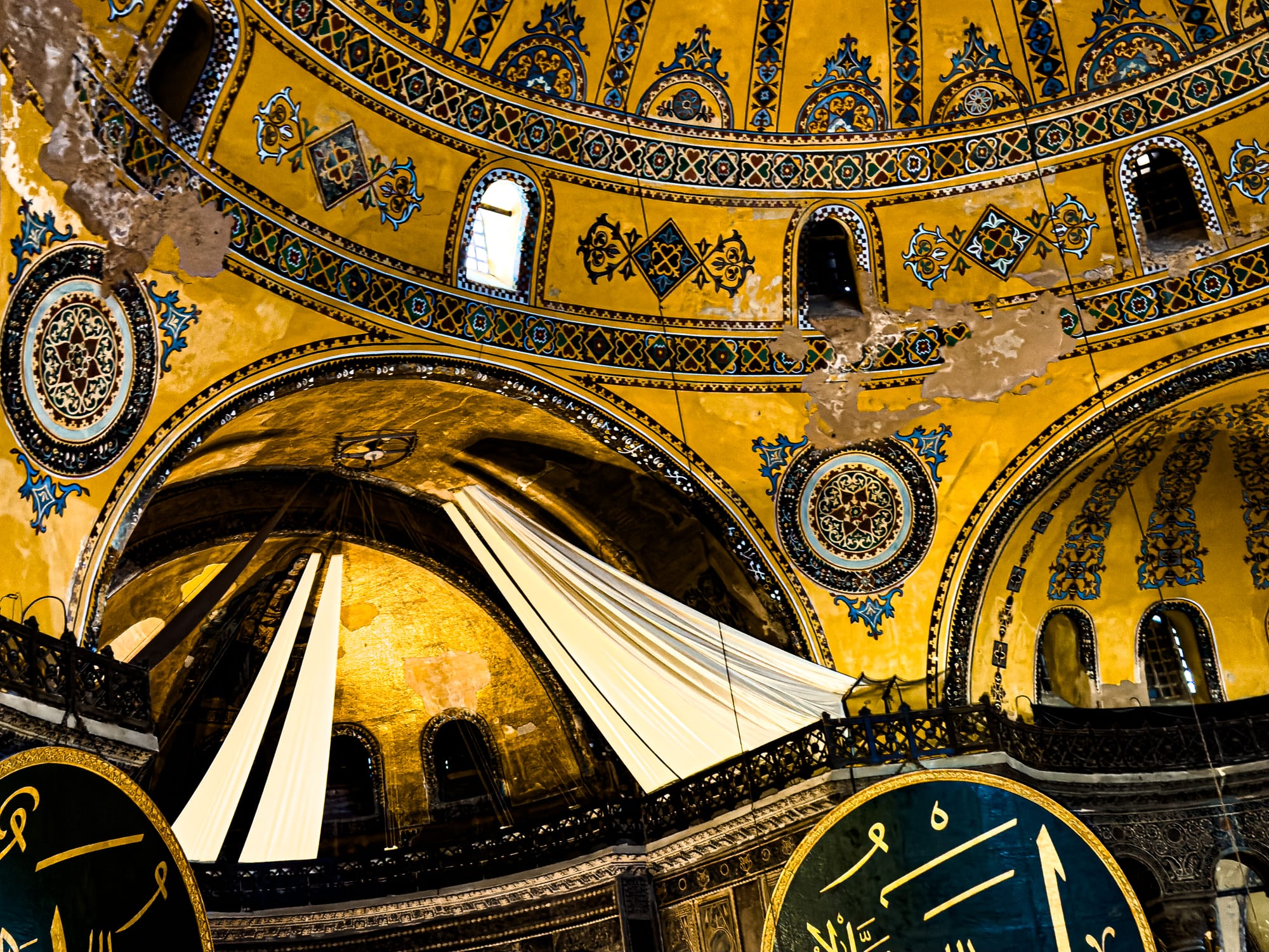 Golden interior of the Hagia Sophia’s dome with Islamic calligraphy medallions, sunlight streaming across faded Christian mosaics, and detailed geometric and floral motifs in blue and gold