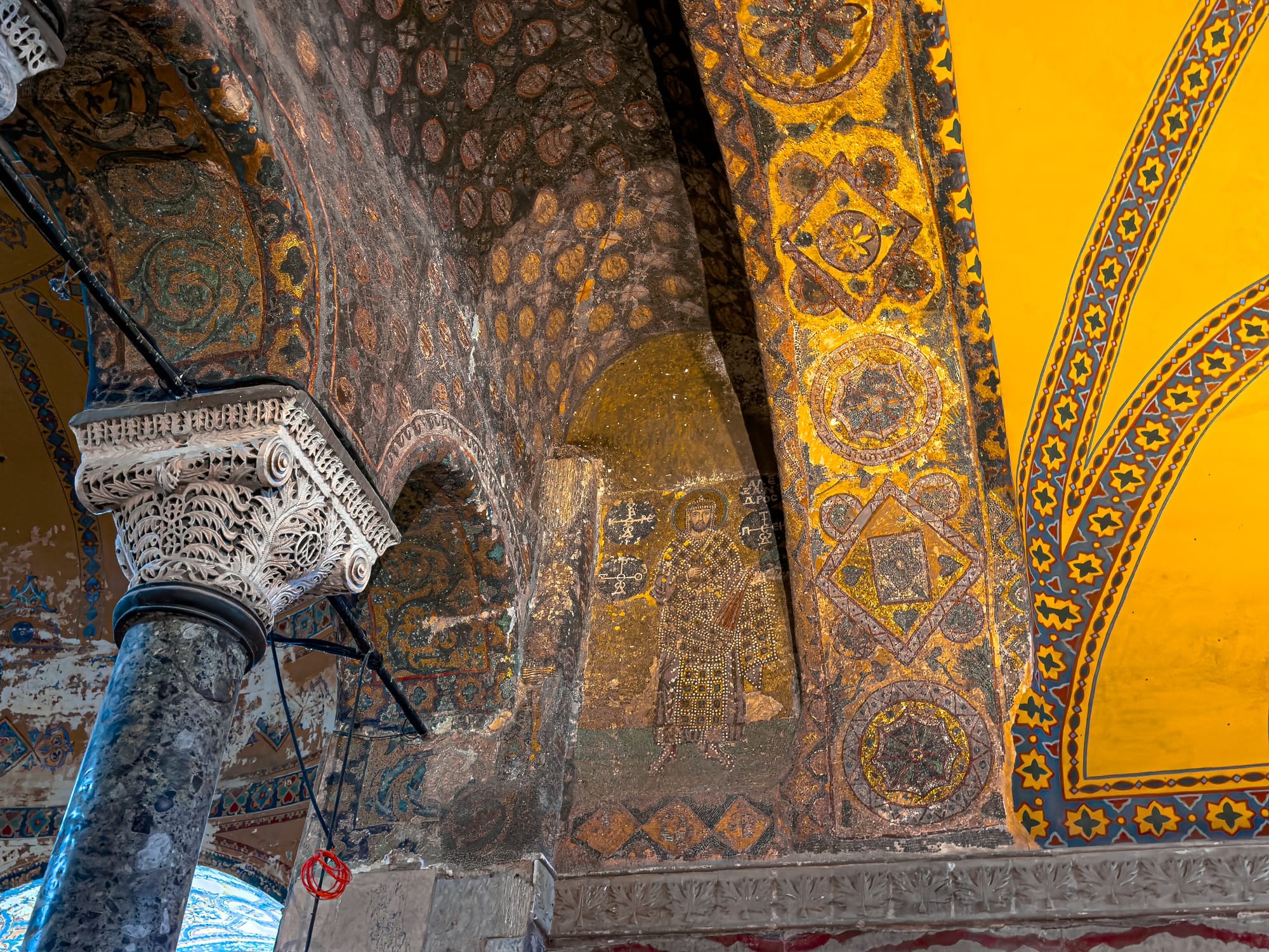 Detailed view of a richly decorated archway in the Hagia Sophia, with faded gold mosaic depicting a Byzantine figure next to intricate stone columns and Islamic geometric patterns