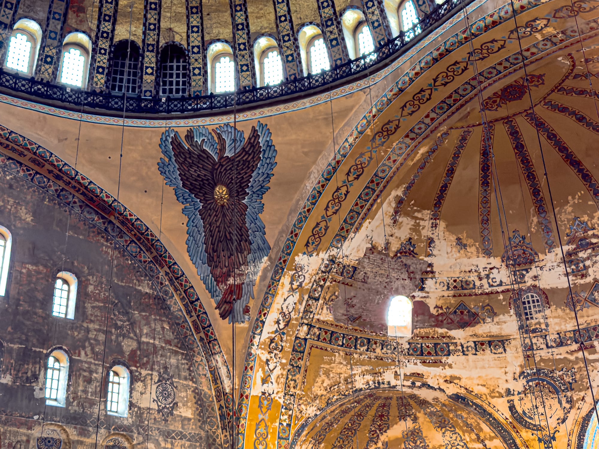 Interior detail of the Hagia Sophia showing a painted six-winged seraphim on the dome ceiling surrounded by ornate golden and blue geometric patterns and partially weathered plaster