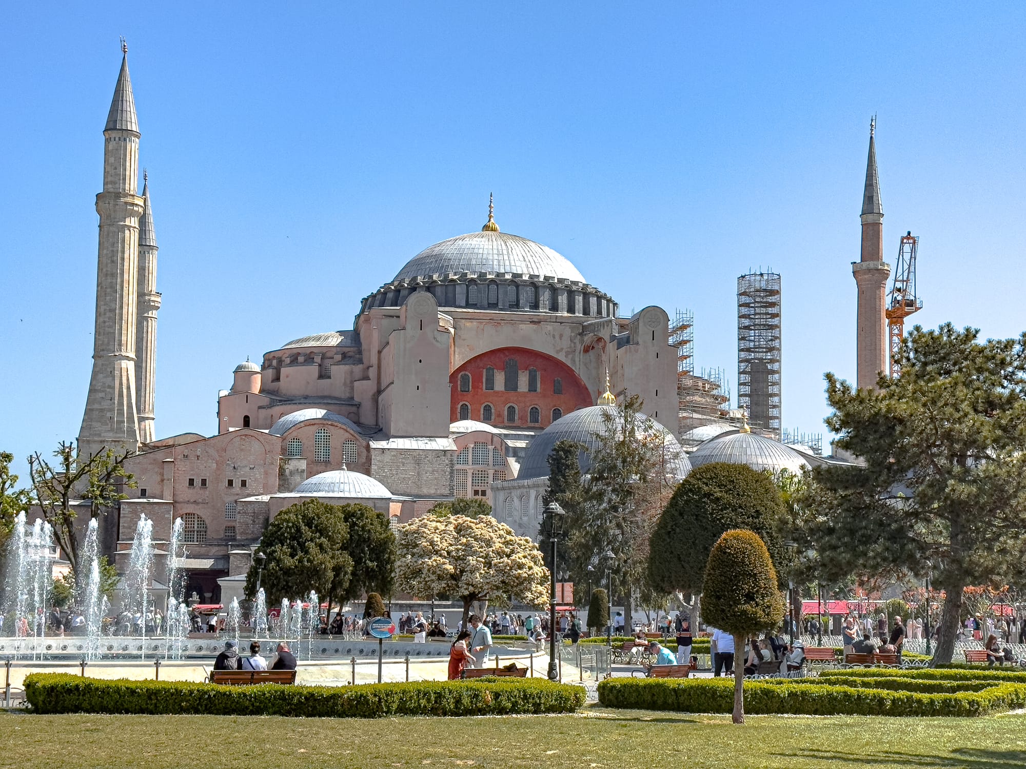 Wide view of the Hagia Sophia’s grand dome and minarets from across the park, with fountains, trimmed hedges, and people sitting on benches in the foreground