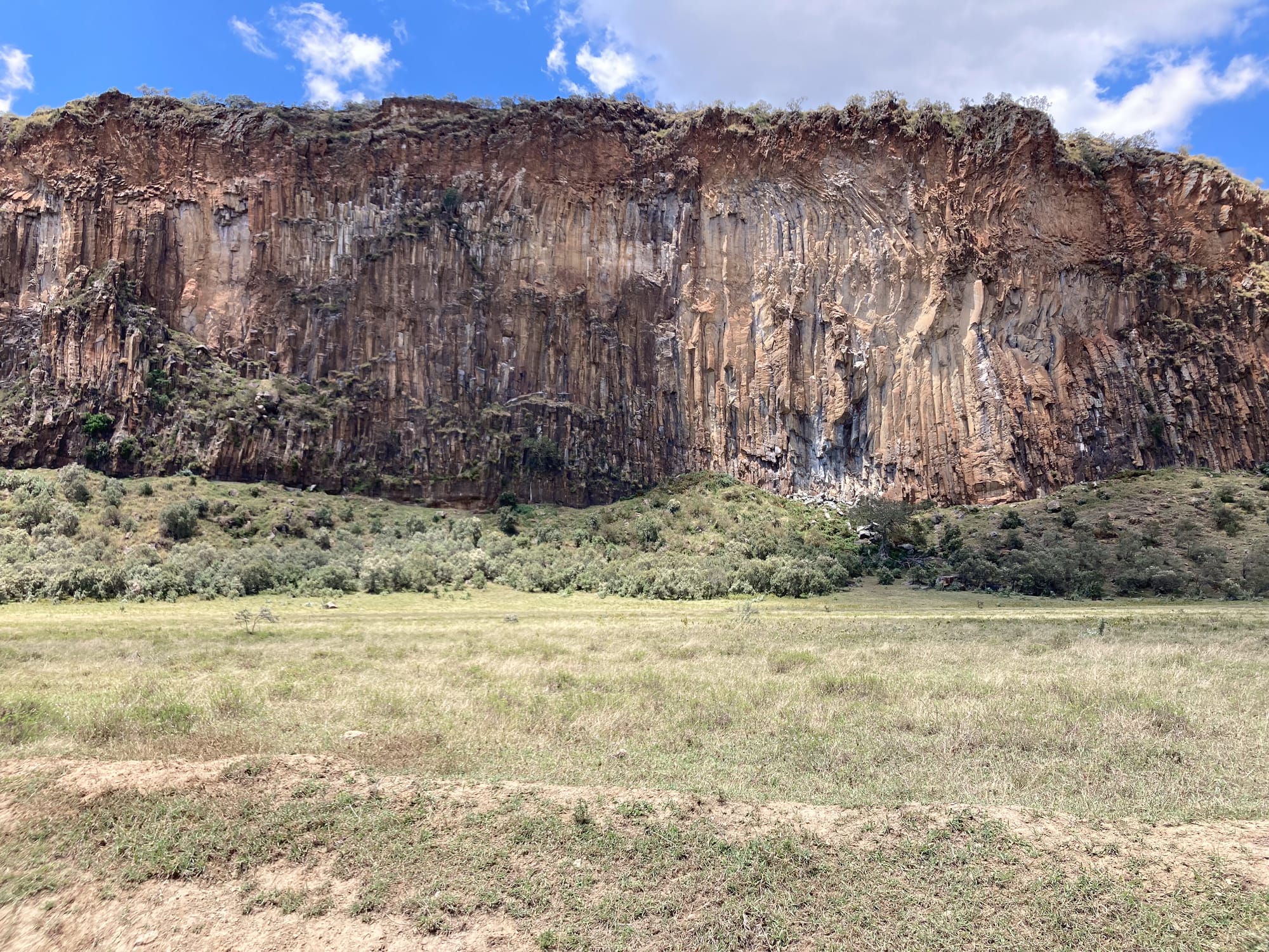 A wide view of a tall volcanic cliff in Hell’s Gate National Park, with vertical rock columns and eroded textures rising above green shrubs and open grassland under a blue sky with scattered clouds