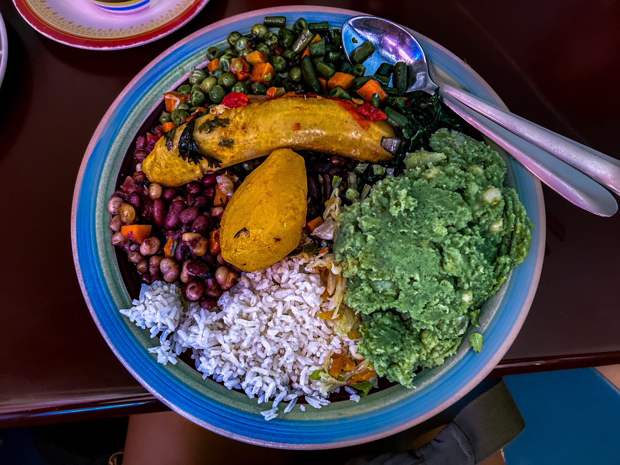 A colorful vegan meal served in a large bowl, featuring rice, beans, sautéed vegetables, greens, mashed potato, and fried banana