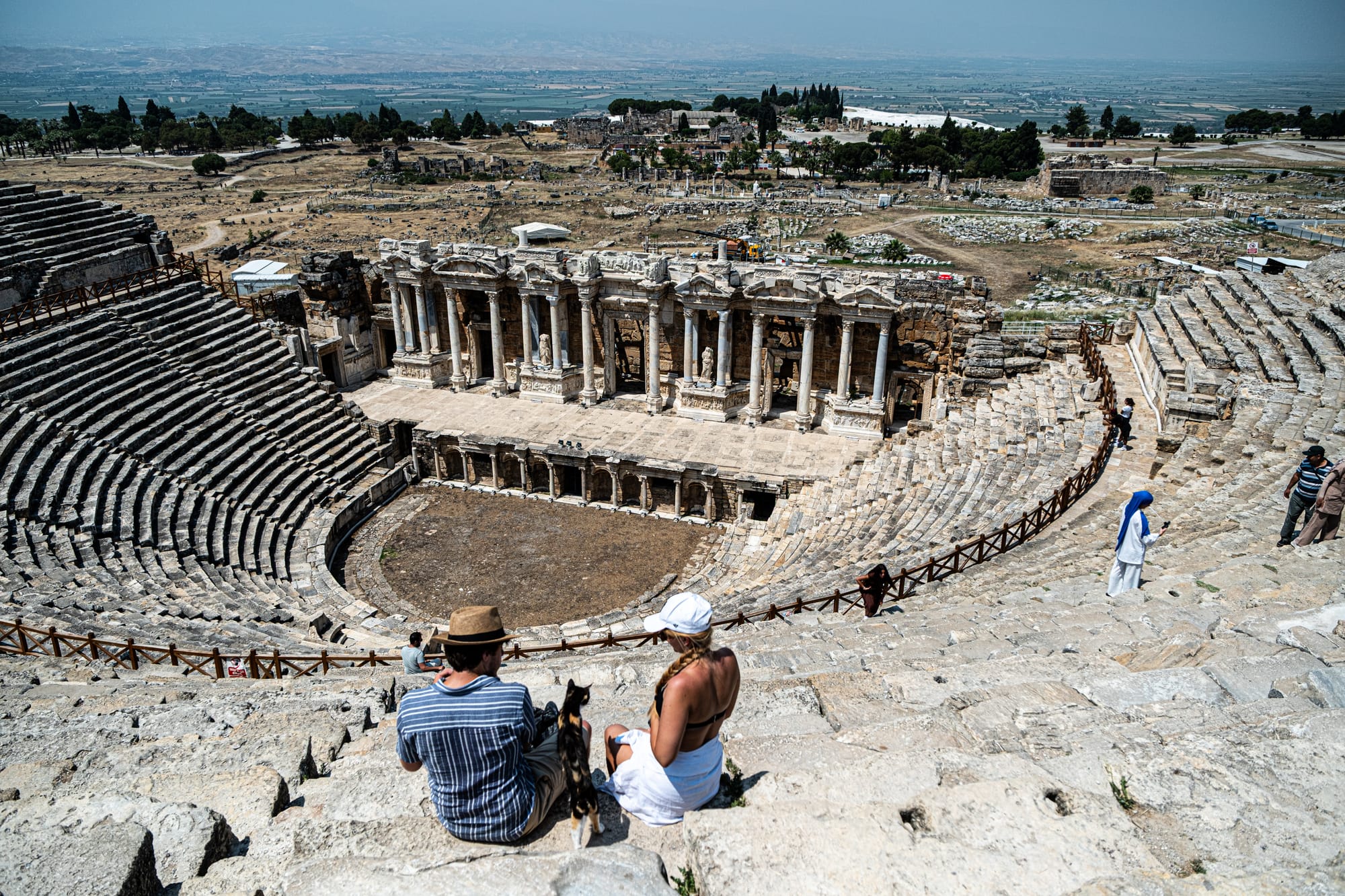 Two visitors, one holding a cat, sit among the stone seats of the ancient amphitheater at Hierapolis, with dramatic ruins and distant countryside in the background