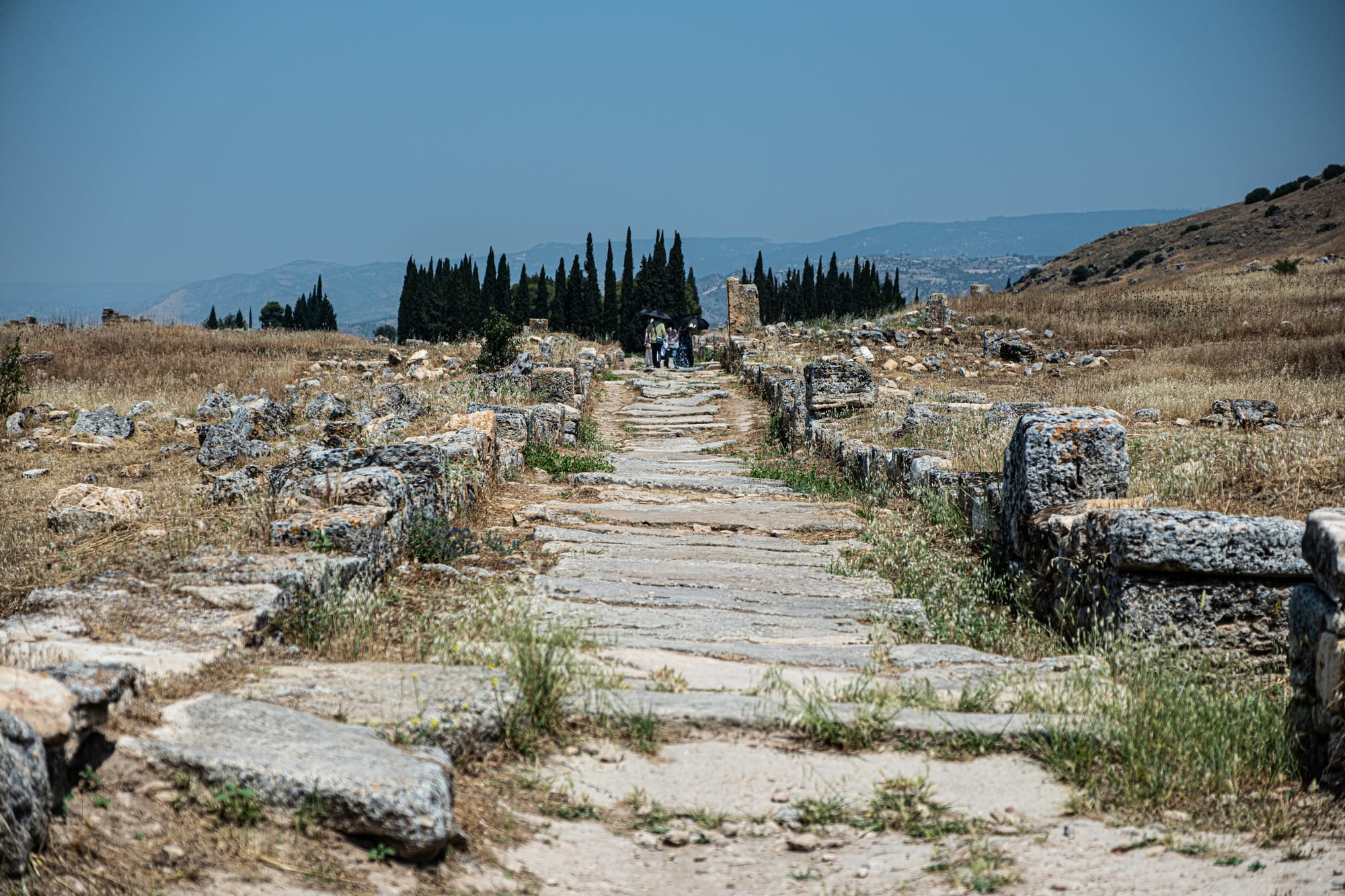 Ancient stone-paved road at Hierapolis, flanked by weathered ruins and leading toward a row of cypress trees in the distance