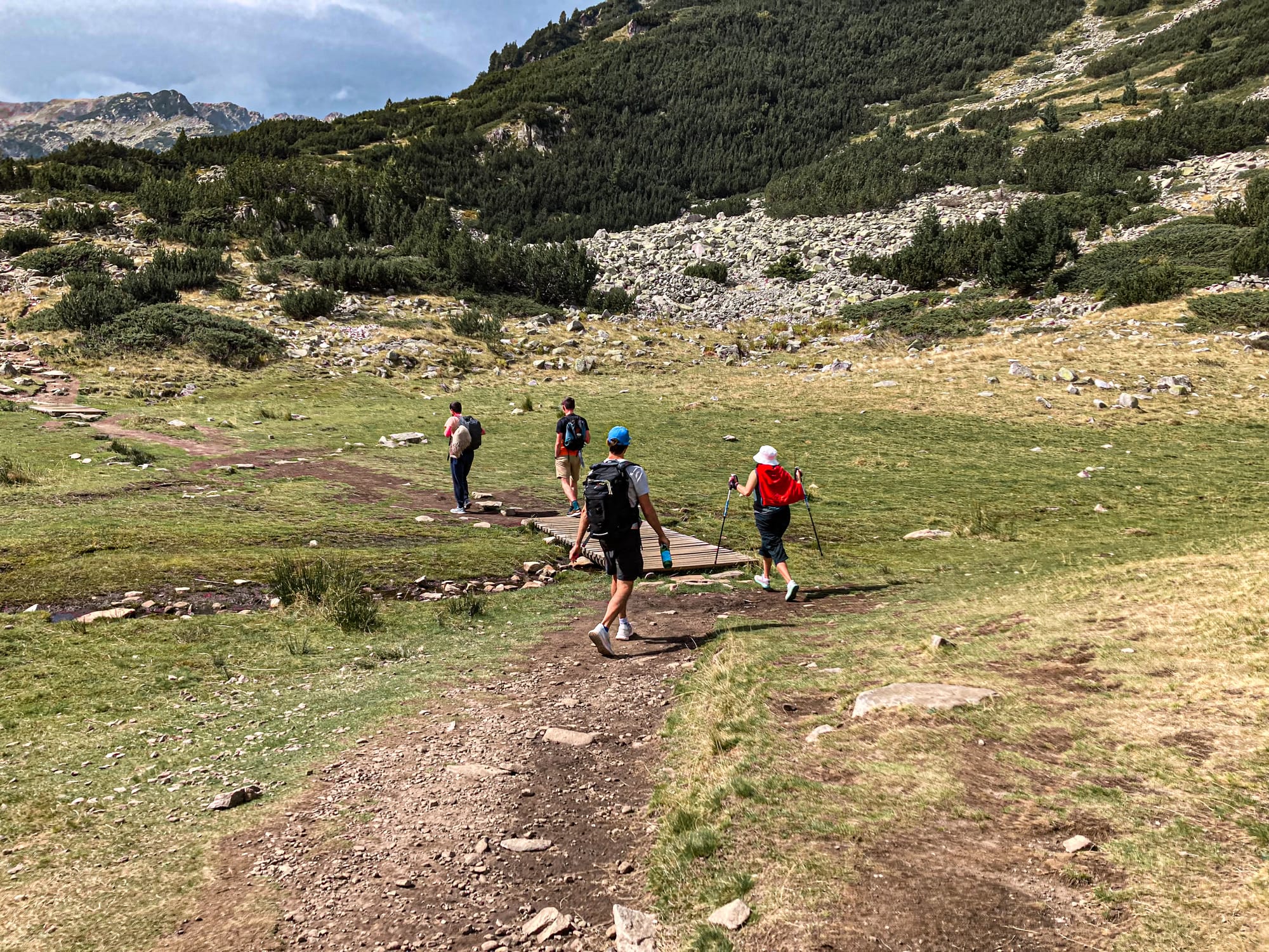 Group of hikers walking along a dirt and wooden trail through a wide alpine meadow in the Pirin Mountains, with rocky slopes and green ridges rising in the background under a partly cloudy sky