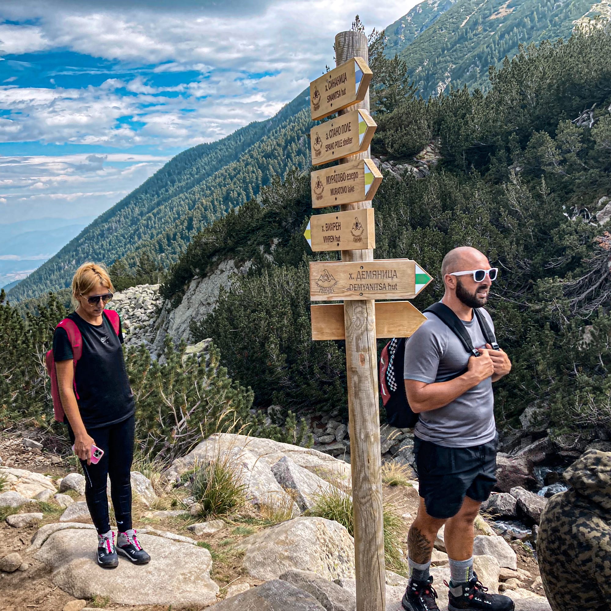 Two hikers standing beside a wooden trail signpost in the Pirin Mountains, with arrows pointing to destinations such as Demyanitsa Hut, Muratovo Lake, and Vihren Peak, set against a backdrop of pine forest and rugged alpine slopes