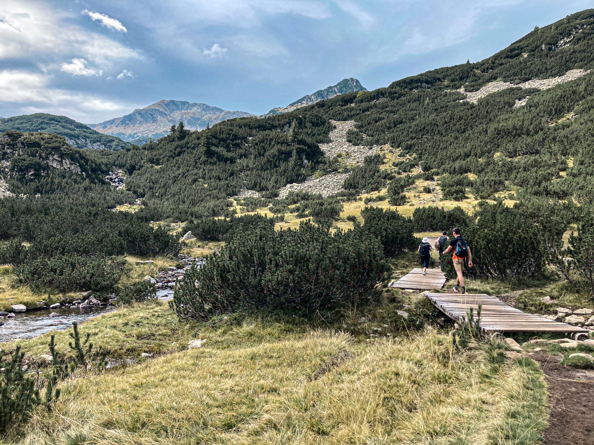 Hikers walking along a wooden boardwalk trail through alpine meadows in the Pirin Mountains, with a stream running alongside and rugged peaks rising in the distance under a cloudy sky