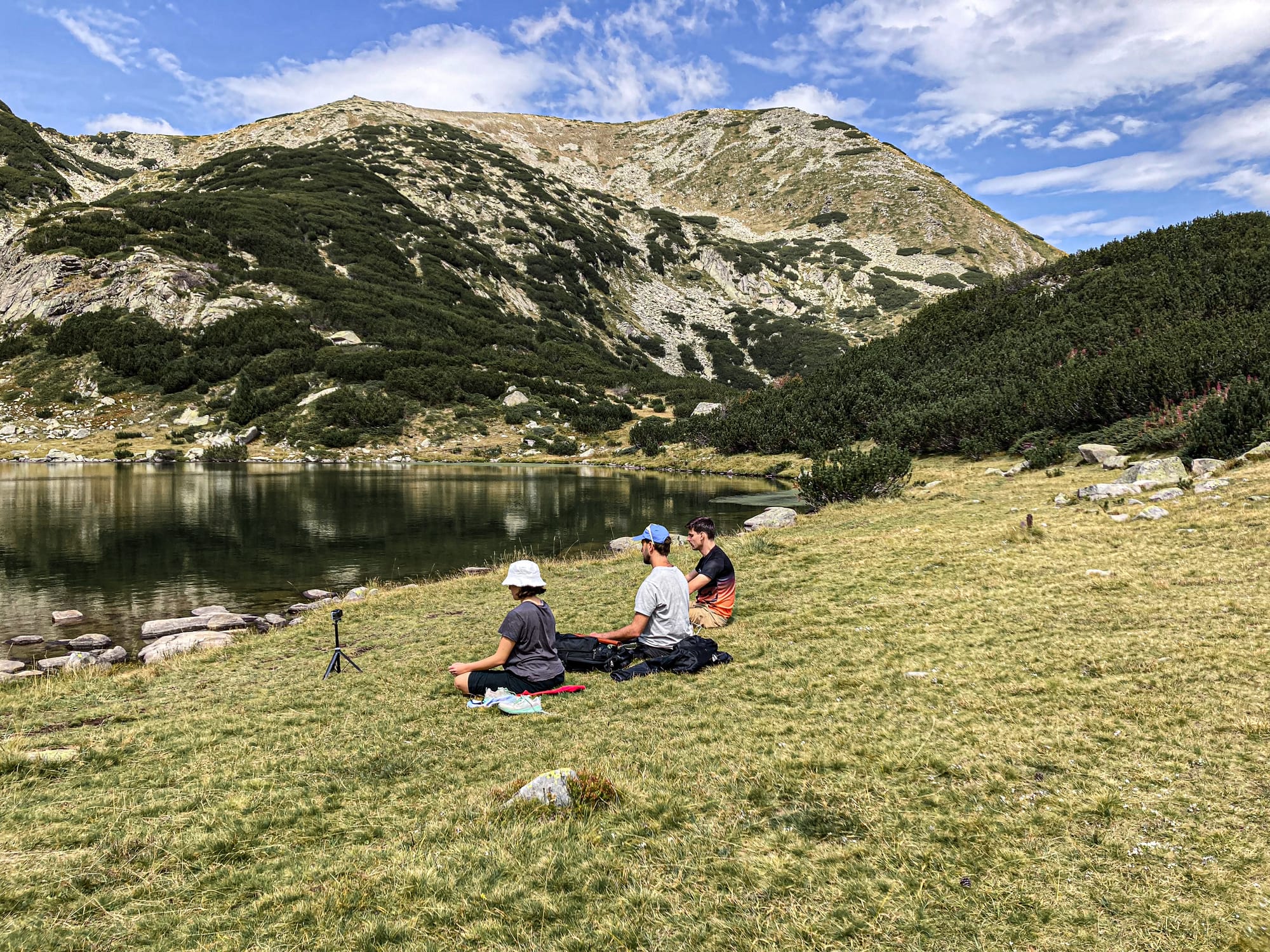 Group of hikers sitting on grassy shores beside a glacial lake in the Pirin Mountains, with clear water reflecting rugged slopes and a partly cloudy sky above