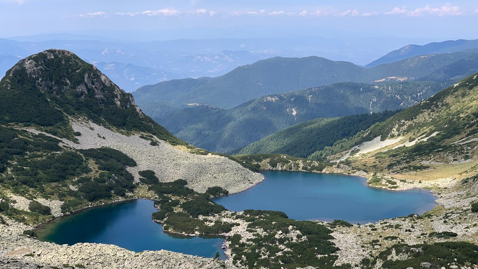 Two glacial lakes in the Pirin Mountains, their deep blue waters surrounded by rocky slopes and green vegetation, with layers of forested ridges stretching into the hazy horizon under a soft summer sky