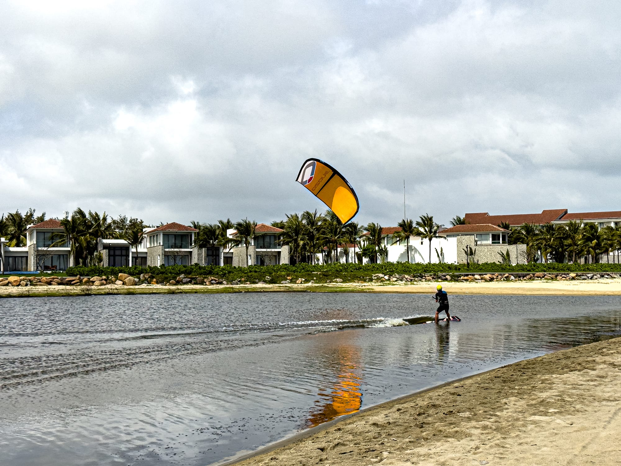 A person kitesurfing with a yellow kite on a lagoon near Hội An, with modern villas and palm trees lining the shore in the background