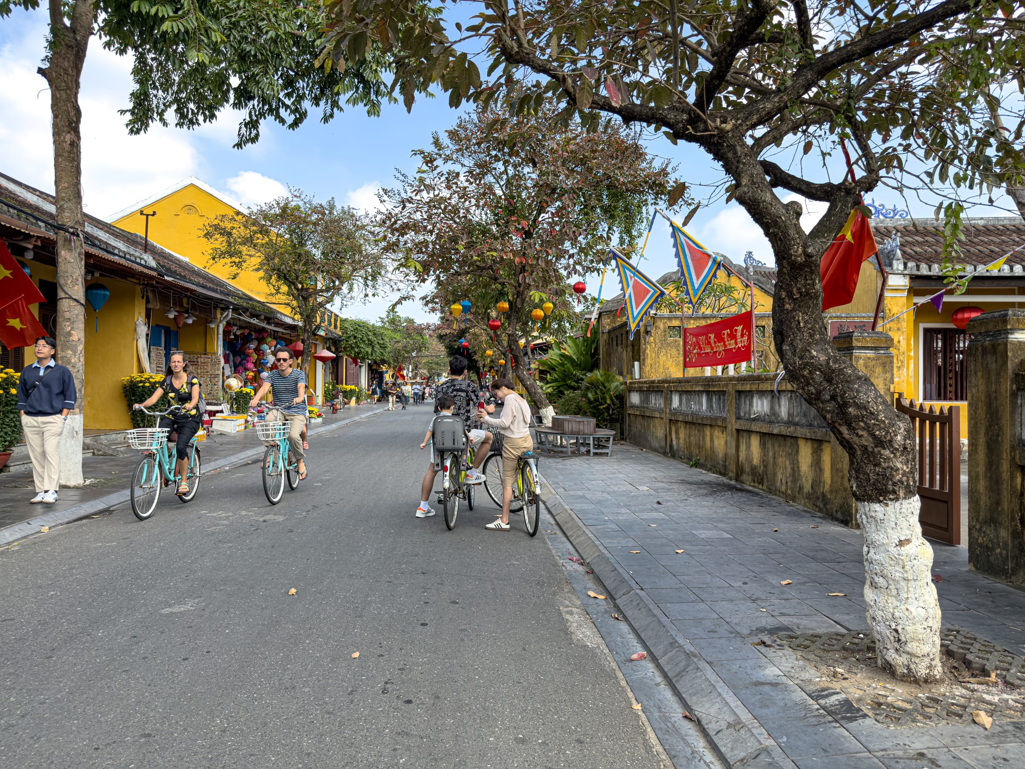 Cyclists riding through Hội An Old Town with yellow shopfronts, lanterns, and tree-lined streets