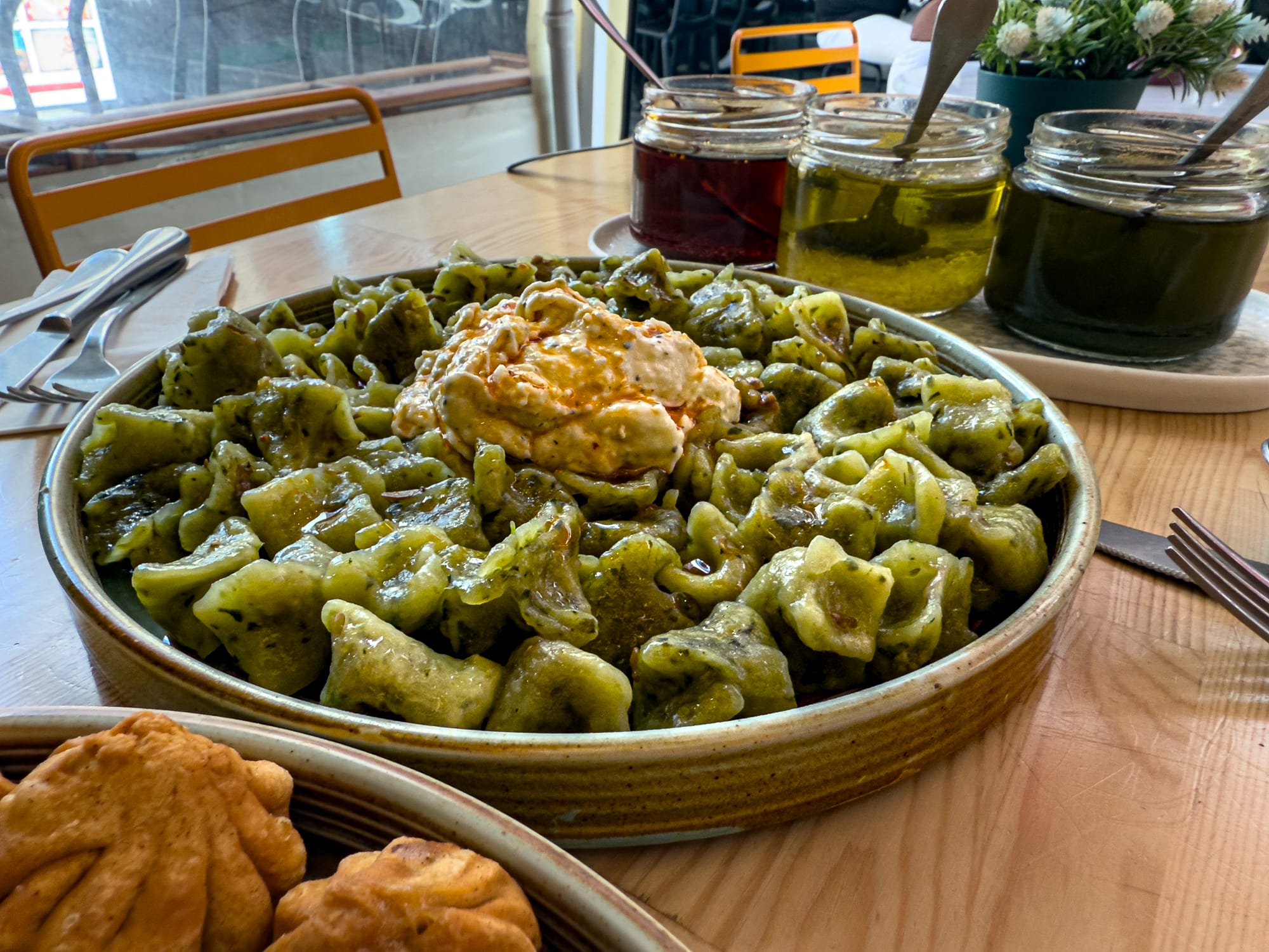 Close-up of a large bowl of vegan spinach mantı topped with spiced yogurt, with condiment jars and utensils in the background