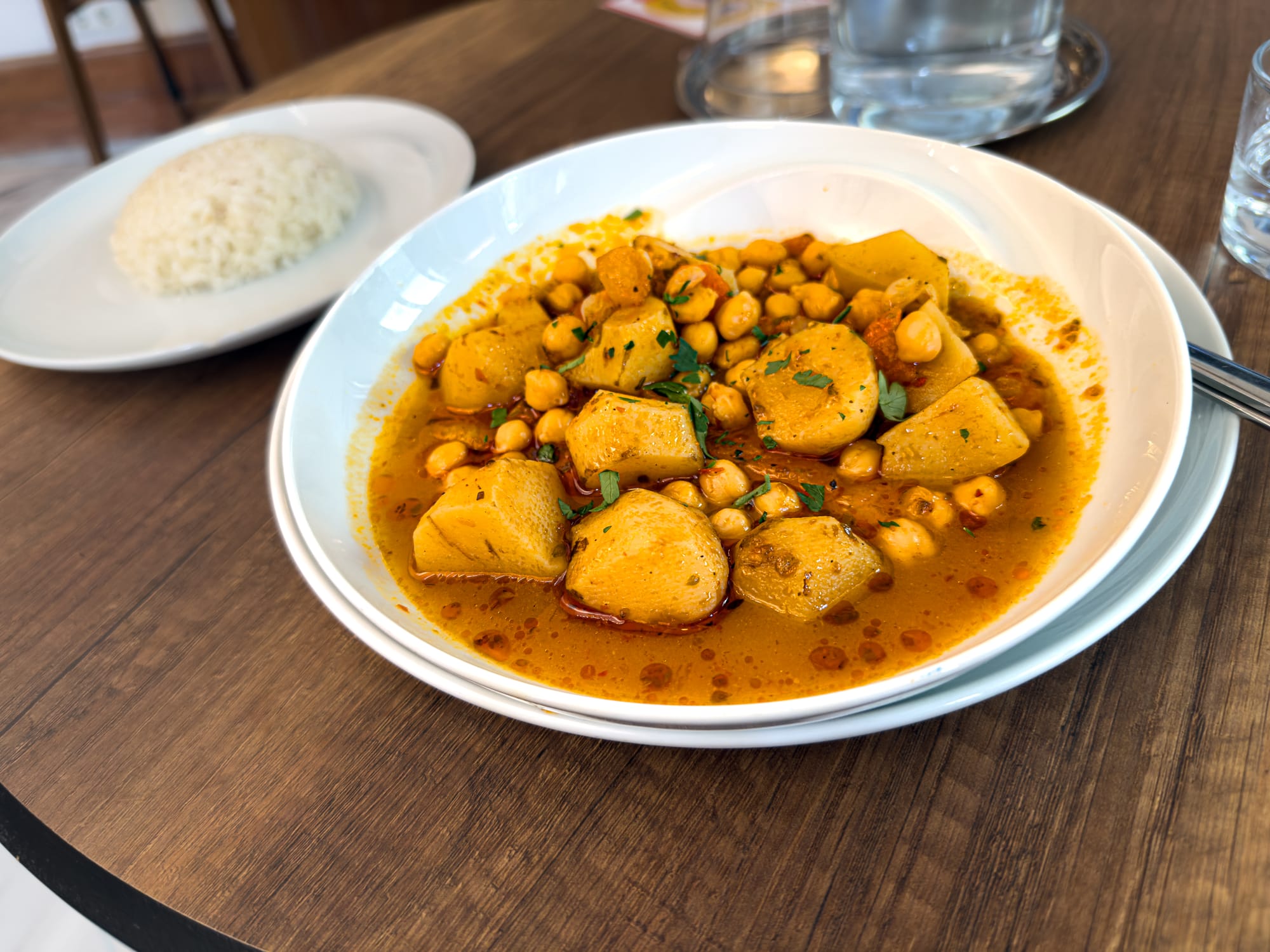 Close-up of a white bowl filled with a Turkish-style sunchoke and chickpea stew in tomato broth, served with a side of white rice on a wooden table