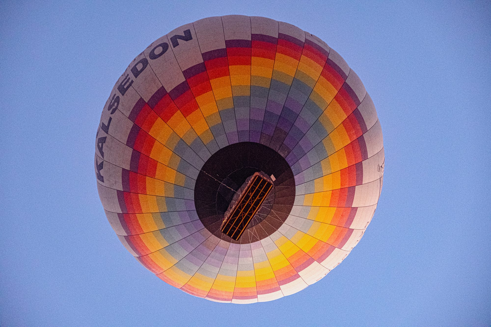 View from below of a rainbow-colored hot air balloon labeled “Kalsedon,” with its basket visible, floating in a clear blue morning sky