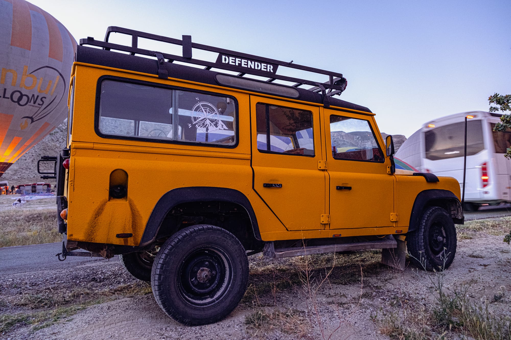 A bright yellow Land Rover Defender jeep with a roof rack parked on the roadside at dawn in Cappadocia, with a partially inflated hot air balloon in the background and another vehicle passing by